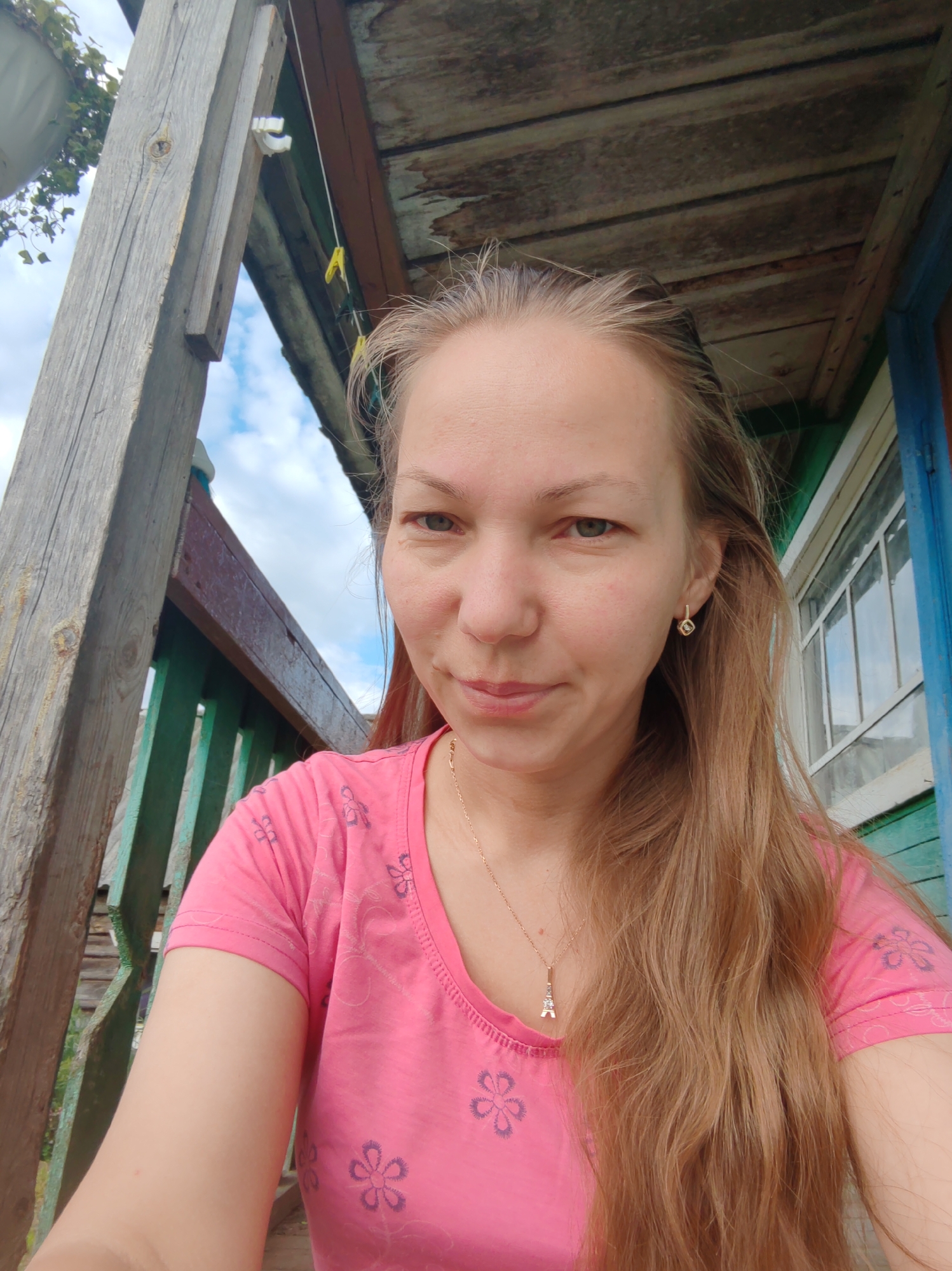 A young woman with long, blonde hair is taking a selfie on a wooden porch. She is wearing a pink t-shirt with a floral pattern and a gold necklace with a small Eiffel Tower pendant. She is also wearing a small gold earring in her right ear. The background shows a wooden structure with a corrugated roof, a green railing, and a window. The sky with clouds is visible in the distance. The setting appears to be a rural or semi-rural location, possibly a dacha or a house in a village. The specific location in the city of Чухлэм, Russia, is not discernible from the image.