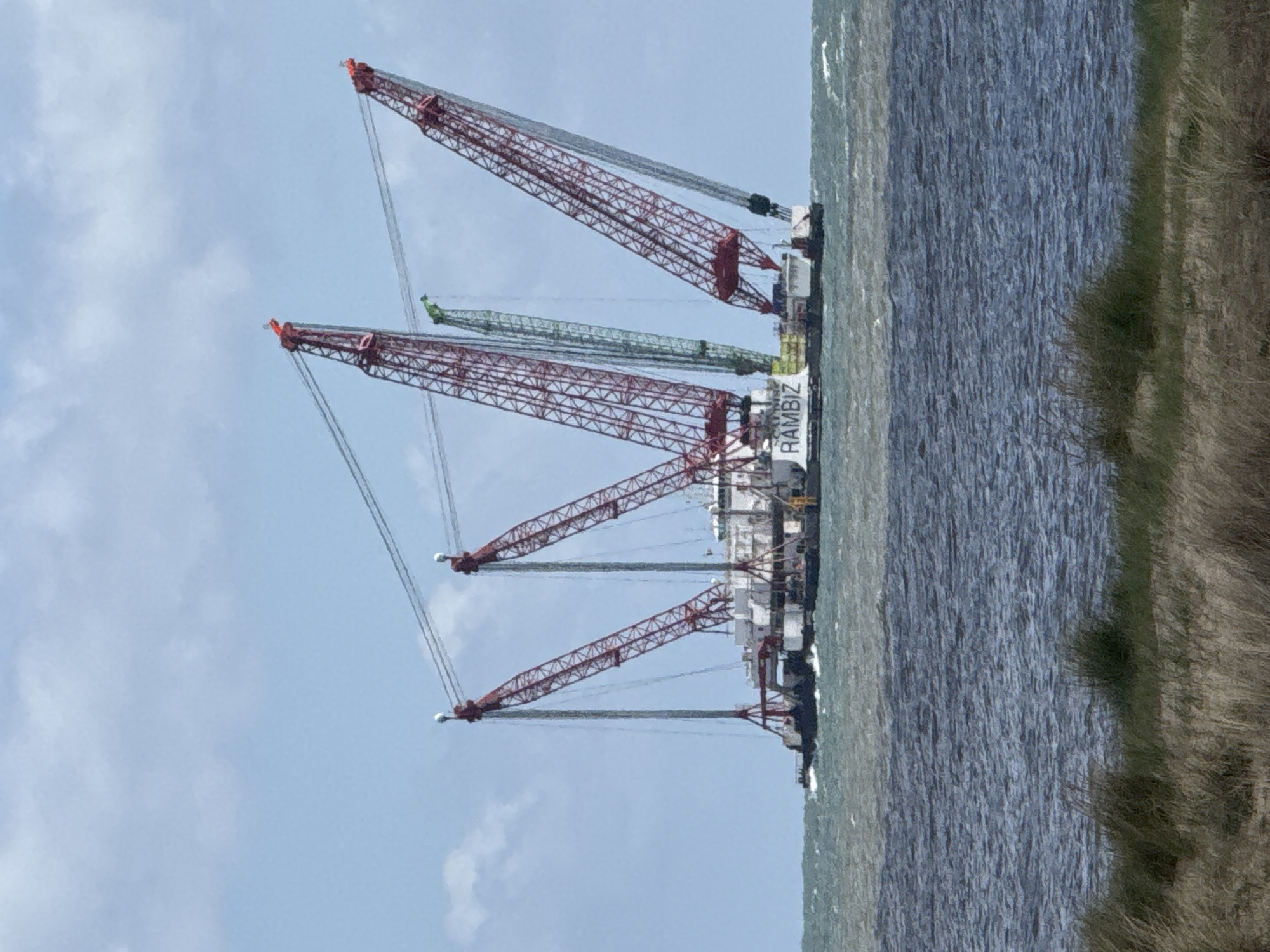 The image shows a large construction barge named "RAMBIZ" with multiple cranes on the water, presumably in Great Yarmouth, United Kingdom. Three large, red lattice boom cranes with cables are prominently visible. There is also a green crane behind them. The barge is positioned close to the shore, where patches of green vegetation can be seen. The sky above is pale blue with scattered clouds, and the water is choppy.