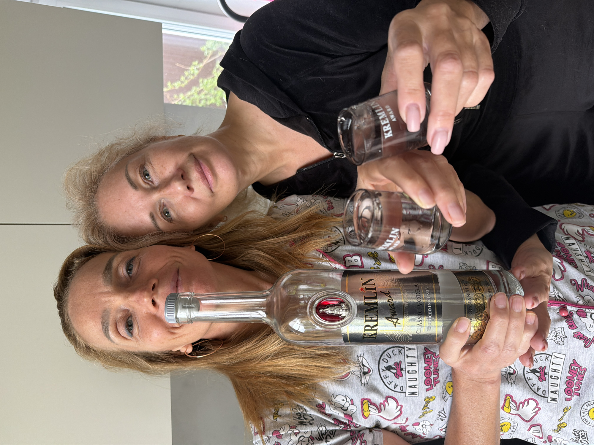 Two women are posing for a photograph, holding a bottle of "Kremlin Award Classic Vodka" and two shot glasses. The bottle's label indicates it is produced in Russia. One woman wears a black top, the other a grey pajama top patterned with "Looney Tunes" characters. They both have fair skin and light-colored hair. The location is not specified as Steklyannyi, Russia. The setting appears to be indoors.