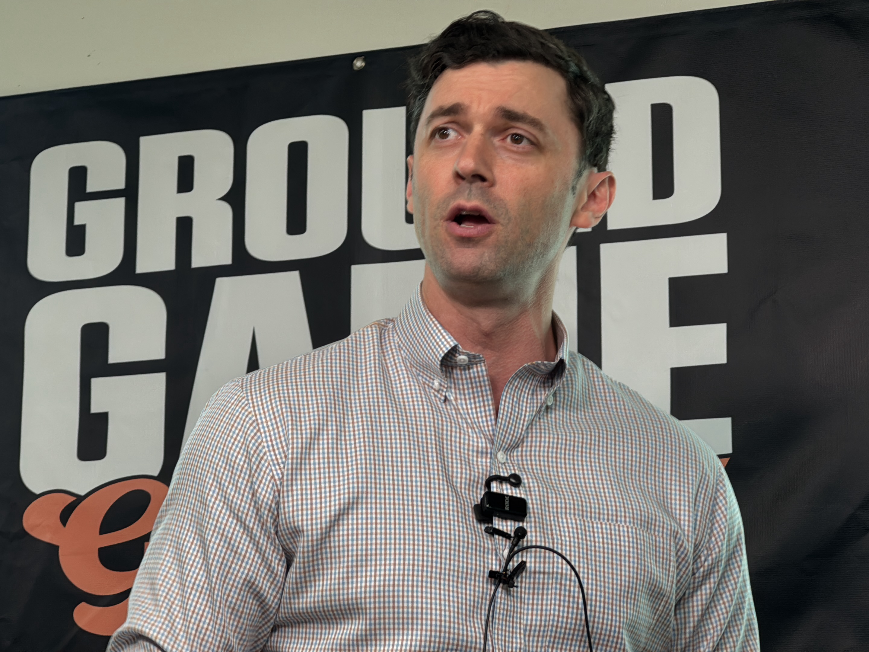 A medium shot captures a man in a button-down shirt, standing in front of a black banner that reads "GROUND GAME" in bold white letters. The shirt is patterned with small brown and blue squares. A microphone is clipped to the front of his shirt. The man is gazing upward and slightly to the right with his mouth open. The background includes a partial view of the "e" in game, and a black ceiling. The picture was taken in the city of Decatur, United States.