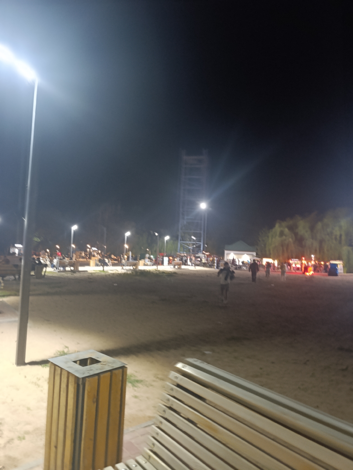The image depicts an outdoor scene at night in Bosteri, Kyrgyzstan. A tall, illuminated lamppost stands prominently in the foreground on the left, casting a bright light. In the middle ground, a wooden trash receptacle with a metal top is visible. Several benches, constructed with metal frames and wooden slats, are partially in view in the foreground and middle ground. The ground appears to be sandy or dirt.
In the background, numerous people are gathered, some seated on benches and others standing. Several streetlights are illuminating the area where the people are congregated. A distinctive tall, metal tower structure is visible in the background, and a small white building with a green roof is also present. Trees with hanging foliage are situated further in the background, suggesting a park or recreational area. The overall impression is of a public gathering space at night.