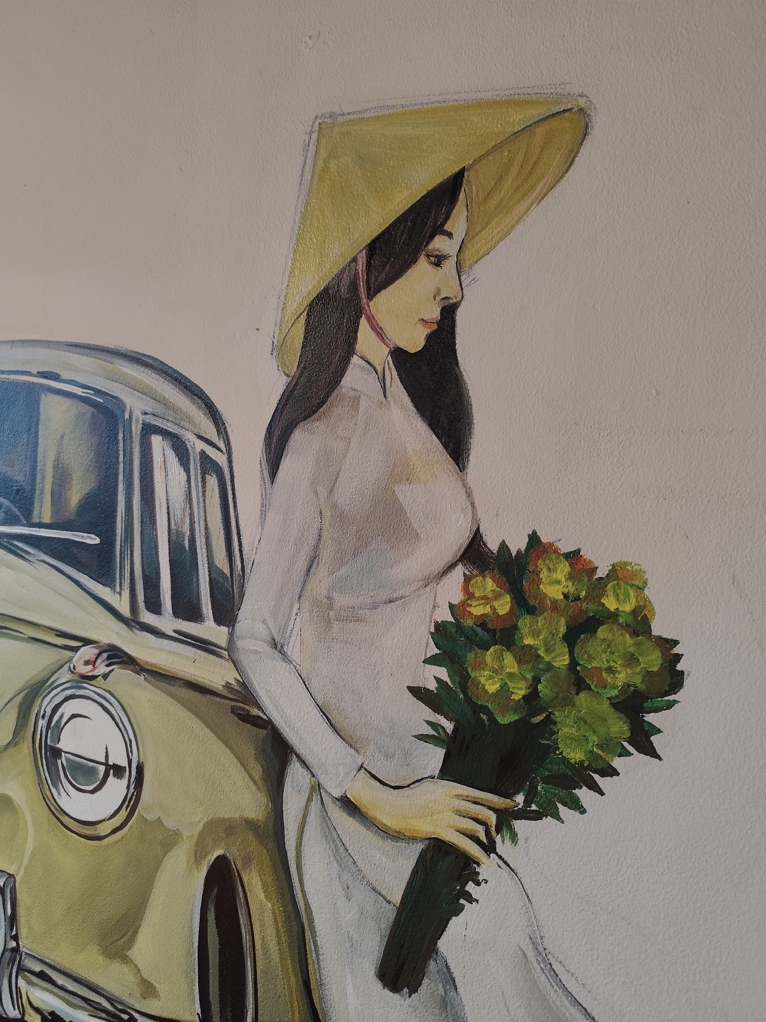 The image depicts a painting on a wall, likely a mural or street art. The subject is a woman wearing a traditional Vietnamese Áo dài (long tunic dress) in white and a conical straw hat (nón lá). She is holding a bouquet of yellow and green flowers. Next to her is a vintage car, likely a European model, painted in shades of green and yellow. The background is a plain wall painted in a light beige color. The style of the painting is somewhat impressionistic. This scene, with the traditional dress, conical hat, and vintage car, is typical of the aesthetic often found in Da Lat, Vietnam, a city known for its French colonial architecture, cool climate, and romantic atmosphere. The painting likely aims to evoke a sense of nostalgia and Vietnamese cultural identity within that specific location.