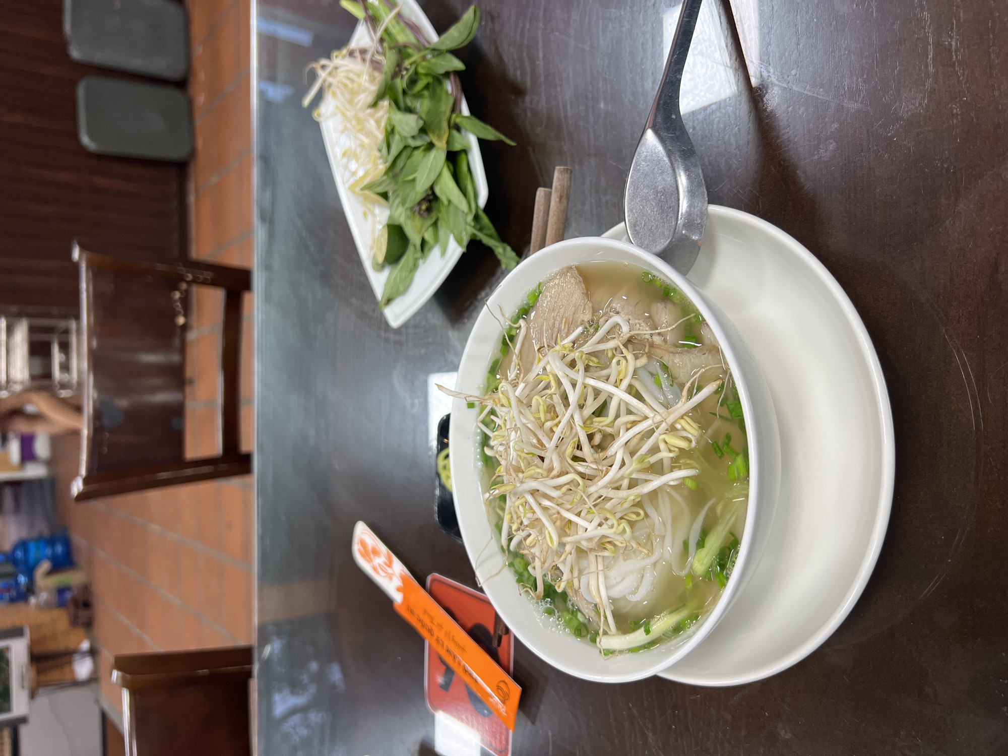The image captures an overhead, close-up view of a bowl of Vietnamese pho, ready to be eaten in what appears to be a restaurant in Bac Nha Trang Ward, Vietnam. The central subject is a large white ceramic bowl filled with pho, featuring a clear broth, white rice noodles, slices of meat (likely beef), and garnished generously with fresh bean sprouts and chopped green onions. The bowl sits on a white saucer.
A metal spoon is resting on the rim of the pho bowl, indicating readiness for dining. To the upper left, a separate white rectangular dish holds a fresh assortment of green herbs, primarily basil leaves, and more bean sprouts, intended as accompaniments for the soup. Next to the pho bowl, a pair of wooden chopsticks rests on a red coaster or stand. An orange paper wrapper for the chopsticks is prominently displayed, featuring Vietnamese text that translates to "HYGIENIC FOOD SAFETY STANDARD" and "HIGH QUALITY", suggesting attention to cleanliness and quality.
The meal is arranged on a dark brown table surface. The blurred background reveals elements of an indoor dining establishment, including the backs of brown wooden chairs and sections of orange brick walls. Further in the distance, out-of-focus shelves or surfaces with various items, including a blue bottle, are faintly visible. The scene evokes the simple pleasure and authentic presentation of a traditional Vietnamese meal.