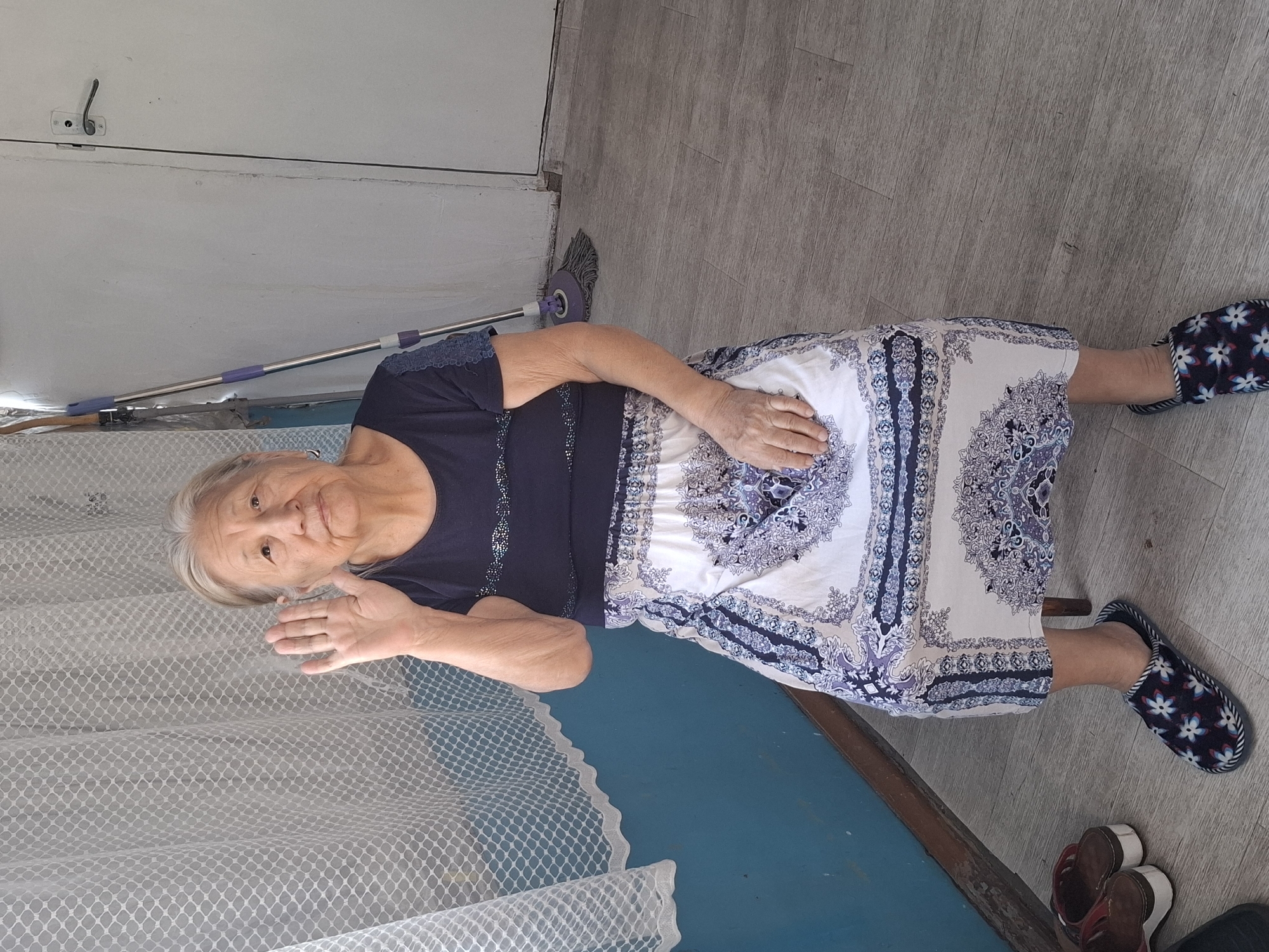 An elderly East Asian woman with grey hair is sitting on a chair, raising her right hand in a waving gesture. She is wearing a dark blue t-shirt with a decorative neckline and a long, patterned skirt with white, blue, and purple designs. She is also wearing patterned slippers. The background consists of a blue wall, a white door with a silver handle, a white lace curtain, and a mop leaning against the wall. A pair of brown sandals are visible on the floor to the left of the woman. The scene appears to be indoors, possibly in a residential setting within Almaty, Kazakhstan. The lighting suggests it is daytime.