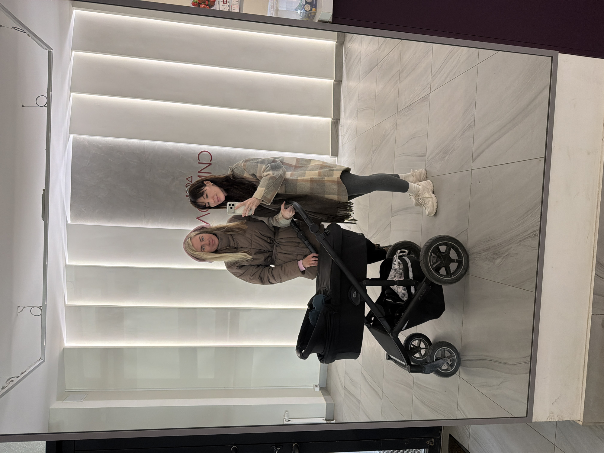 A mirror selfie captures two women and a baby stroller in a modern indoor setting. The image is oriented vertically, with the subjects appearing to be leaning or tilted.
The woman on the left, holding a smartphone to take the picture, has long dark hair and is wearing a plaid jacket, dark leggings, and light-colored sneakers. She has a slight smile. The woman on the right, with blonde hair and wearing a brown puffer jacket and a pink head covering, holds the handle of a dark-colored baby stroller and also smiles gently towards the camera. The stroller appears to be robust, with multiple wheels visible.
The background, reflected in the mirror, features a contemporary interior design. A prominent wall behind the subjects has horizontal panels with integrated lighting, creating illuminated strips. The floor is covered with light-toned, possibly marble-patterned, tiles. In the upper part of the reflection, some reversed and partially visible text, likely "CNDA" or similar, is present on a grey panel, suggesting a brand name or store signage. A shelf or display with items, including what looks like strawberries, is visible in the top right corner of the reflection. The overall environment suggests a commercial space, possibly a mall or a boutique. The scene depicts a casual moment, likely during an outing in Perm, Russia.