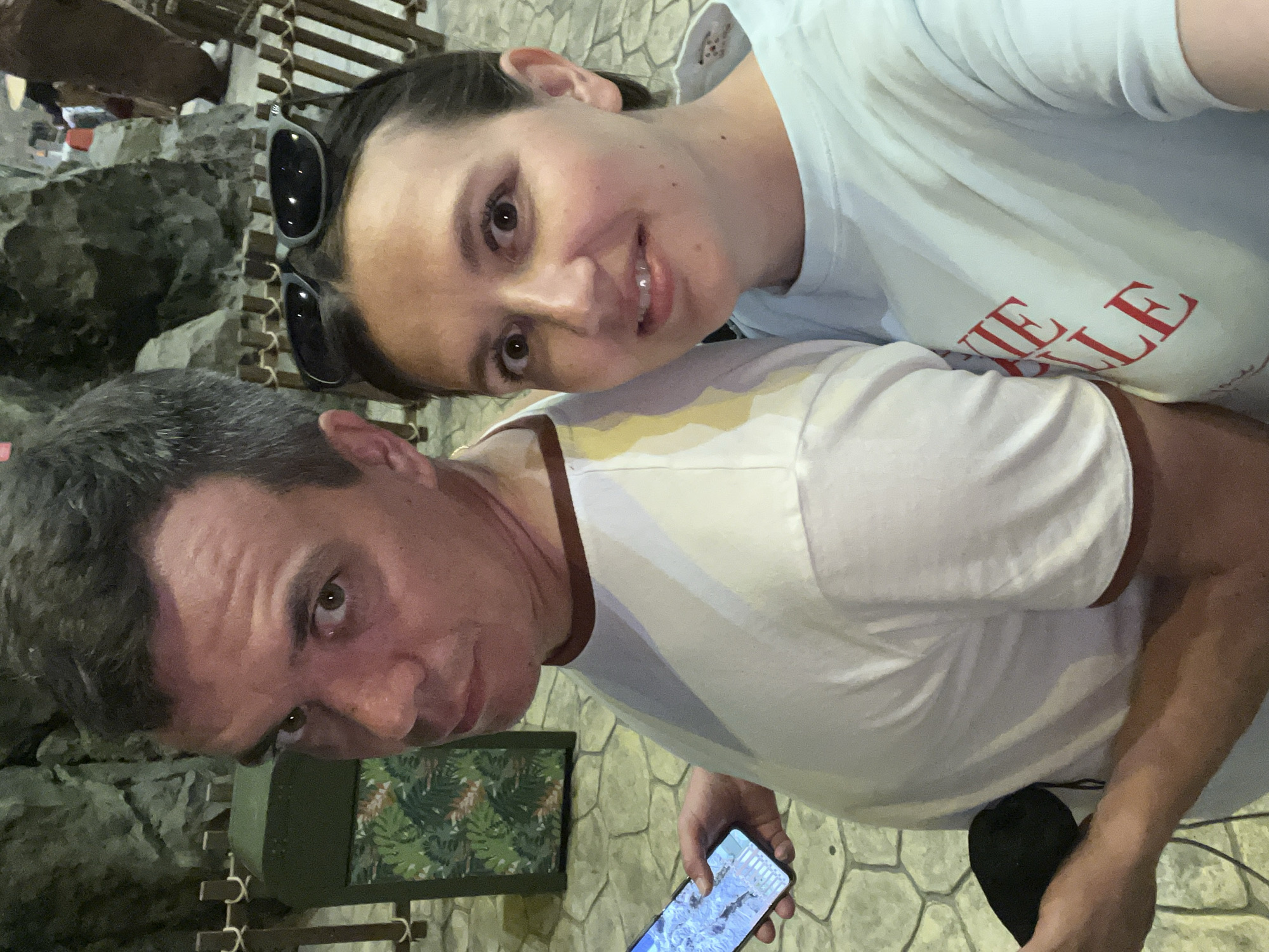 A selfie shows a man and a woman in an indoor environment with stone-like walls, possibly a themed attraction or a museum. The man, on the left, has short, dark hair and is wearing a white t-shirt with a reddish-brown trim. He is holding a smartphone in his left hand, displaying what appears to be a map or navigation app. The woman, on the right, has dark hair and is wearing a light blue t-shirt with red lettering that reads "CHIKIE BALLE". She has sunglasses pushed up on her head. Behind them, a dark green trash receptacle with a tropical leaf pattern is visible, along with a wooden fence and dimly lit rocky structures. The flooring is made of irregularly shaped grey and beige stones. The lighting is dim, suggesting an indoor setting at night or in a low-light area.