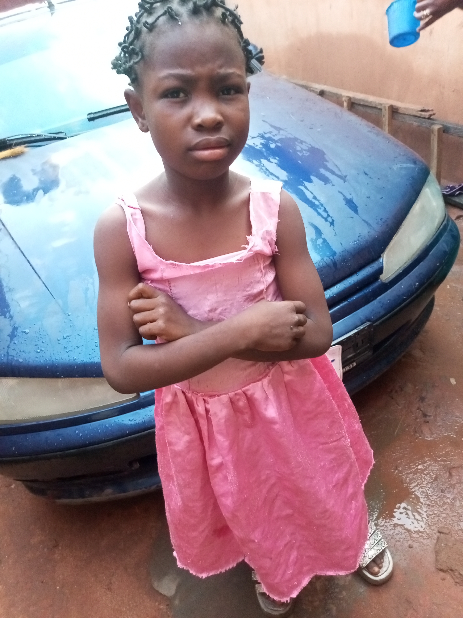An African child stands with arms crossed in front of a blue vehicle. The child is wearing a pink dress and has braided hair. The ground is wet and appears to be dirt. In the background, a person is holding a blue bucket. The scene is located in Tempure, Nigeria.