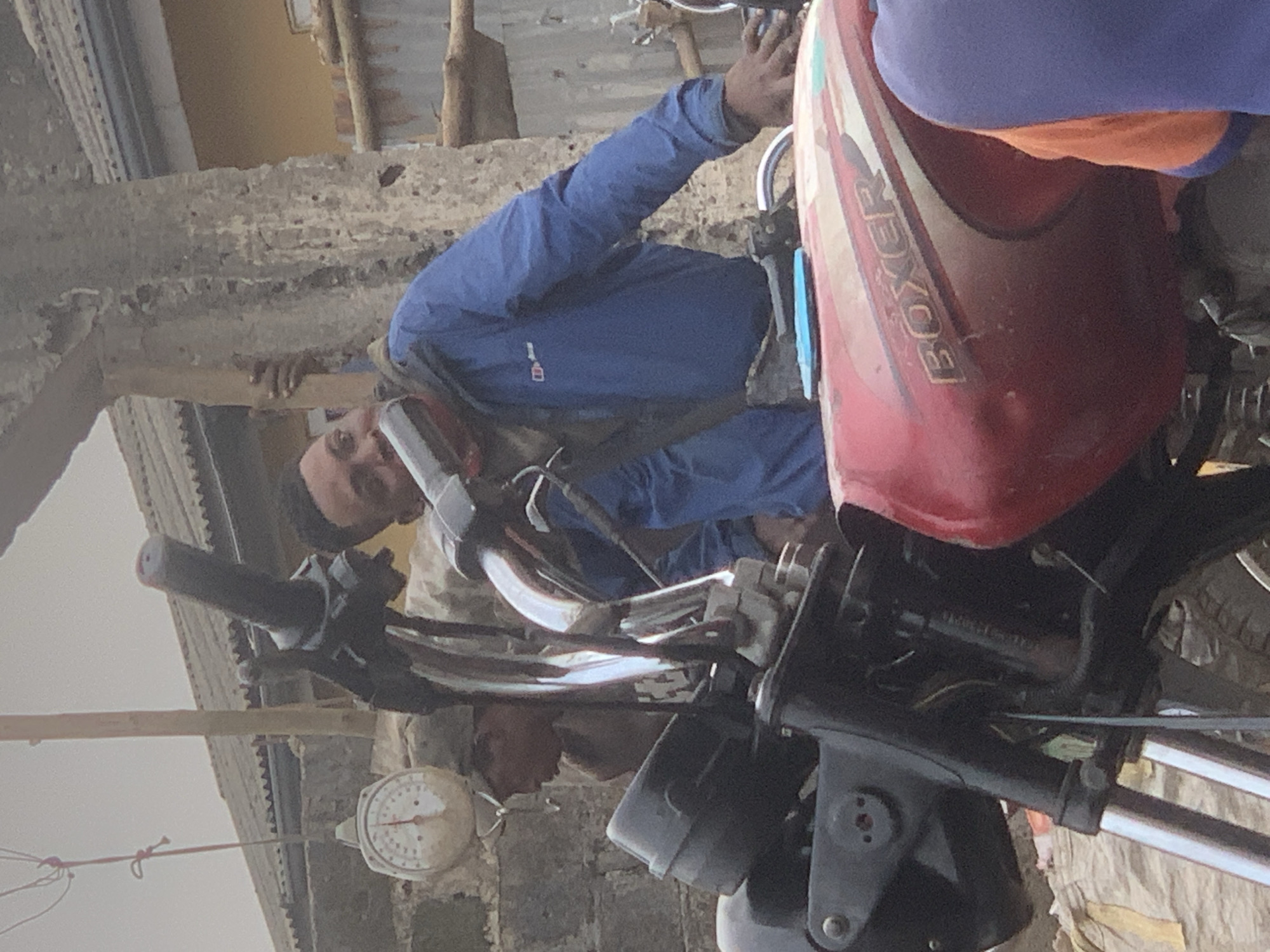 In the city of Shagamu, Nigeria, a man in a blue jacket stands near a red motorcycle. The motorcycle has "BOXER" written on its fuel tank. A scale is visible in the background. The scene appears to be outdoors, possibly in a market or a street.