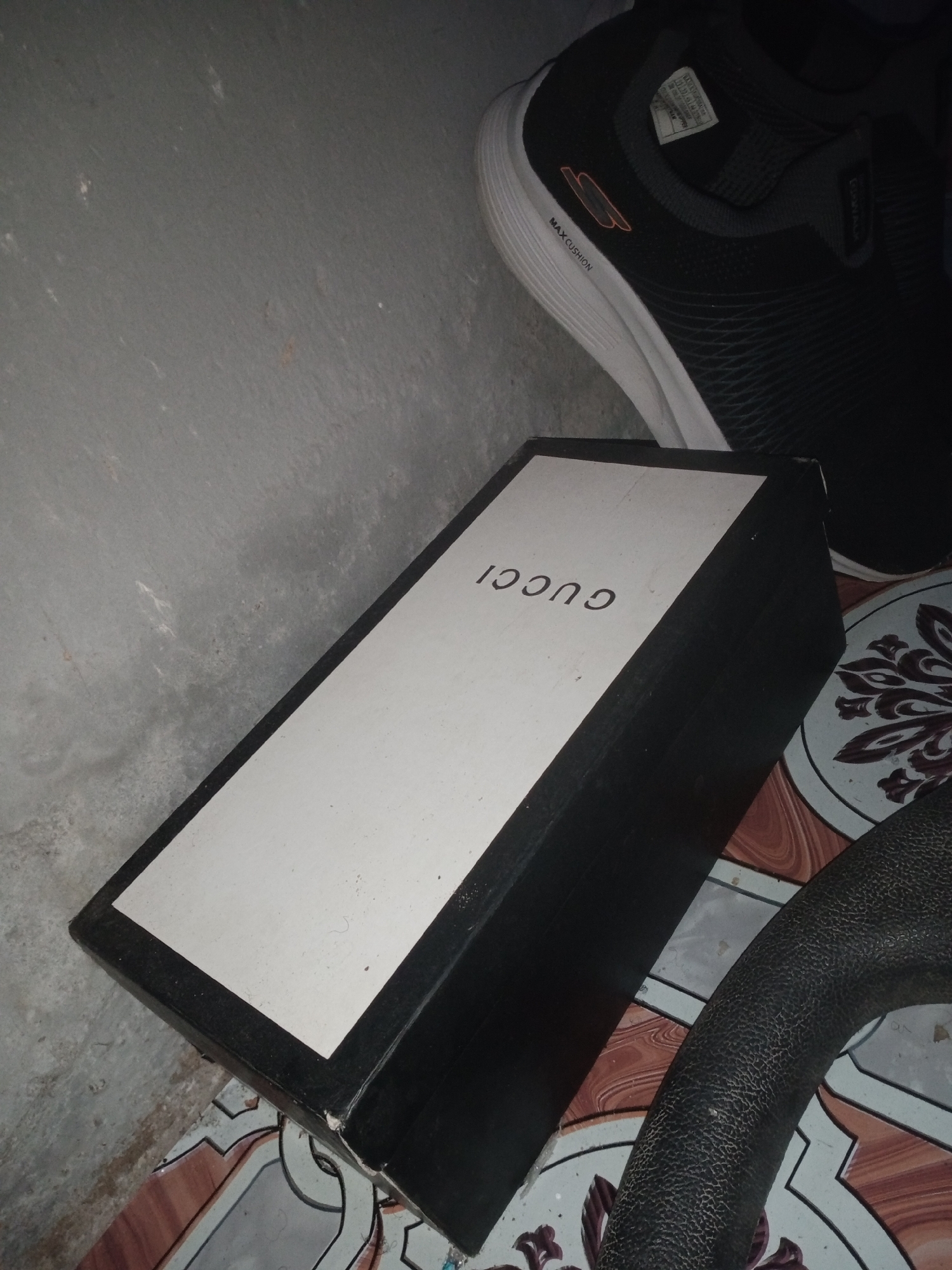 A black Gucci shoe box with a white lid is on the floor in Jalingo, Nigeria. The box is open, revealing the inside. The lid has the word "GUCCI" printed on it in black letters. To the right of the box is a black sneaker with a white sole. The sneaker has "MAX CUSHION" and a logo printed on it. The floor is tiled with a pattern. A grey concrete wall is visible in the background.