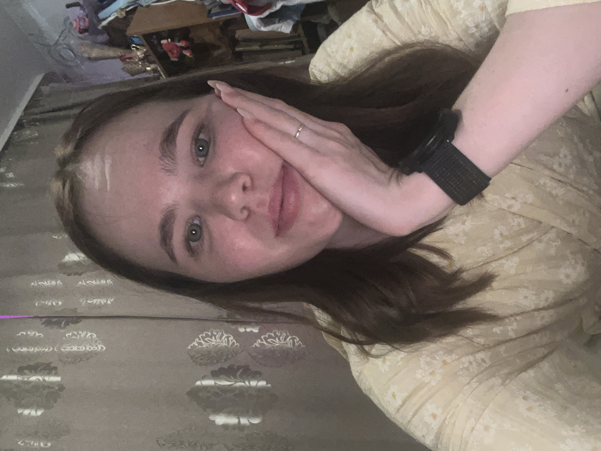 A young woman with fair skin, light brown hair, and blue eyes is shown in a close-up, slightly angled selfie. She has her right hand touching her cheek, revealing a silver ring on her finger. She is wearing a dark-colored watch on her left wrist and a light yellow dress with a small floral print. The background is slightly blurred, showing a patterned brown curtain or wall covering and a cluttered area with boxes and other items. The location in Chaikovsky, Russia is not discernible from the image provided.