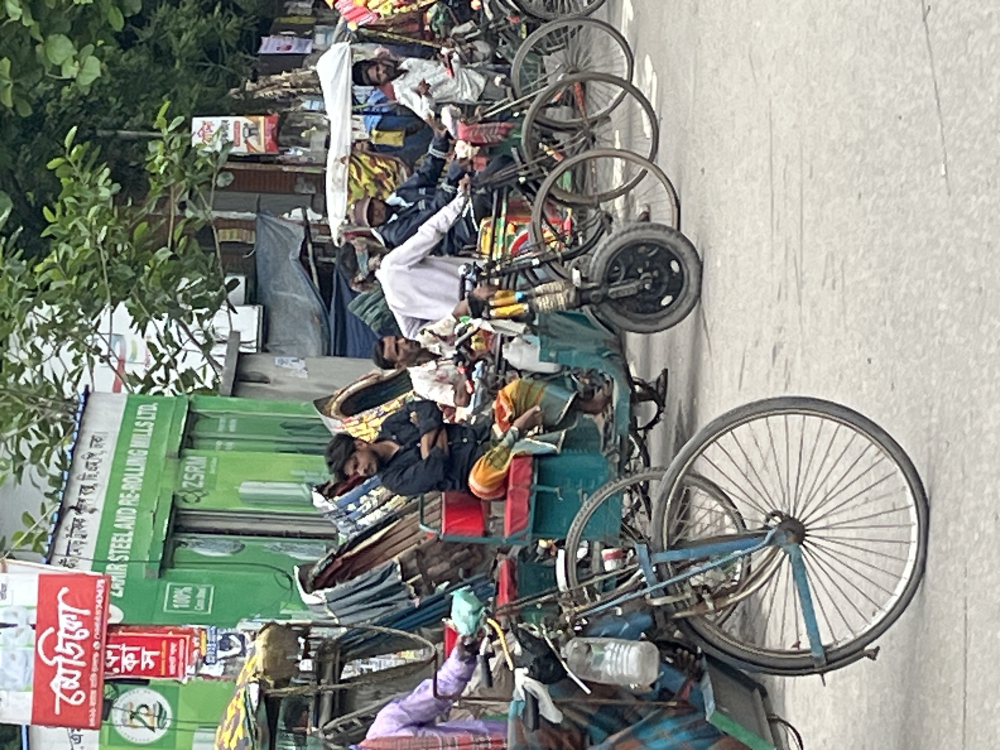 The image captures a bustling street scene in Dhaka, Bangladesh, dominated by parked and occupied rickshaws or three-wheeled passenger vehicles. Several men, likely rickshaw pullers, are visible; some are sitting on their vehicles, while others appear to be resting or sleeping. One man in a dark blue shirt is distinctly seen lying down and resting on the seat of his vehicle, with a colorful cloth wrapped around his legs. Another man in a white shirt and cap is seated on a rickshaw further down the line. The street appears to be paved, and the overall lighting suggests it is daytime with clear weather.
In the background, there is dense greenery from trees and bushes on the left side. Various buildings and stalls line the street, with several signs displaying text.
Notable text visible in the image includes:
- A red sign with "মেডিকো" (Mediko), likely a business name.
- Another sign with "কট মোড় ট্রাফিক পুলিশ বক্স, ছি:এমপি, ঢাকা।" (Kot Mor Traffic Police Box, C.M.P., Dhaka), indicating the specific location as a traffic police box in Dhaka.
- A prominent green signboard displaying "AMIR STEEL AND RE-ROLLING MILLS LTD." and its abbreviation "ZSRM," along with "100%" and "Com B," suggesting a commercial establishment.
The scene depicts a typical urban environment in Bangladesh, characterized by manual and pedal-powered public transport and local businesses, conveying a sense of daily life and work in the city.