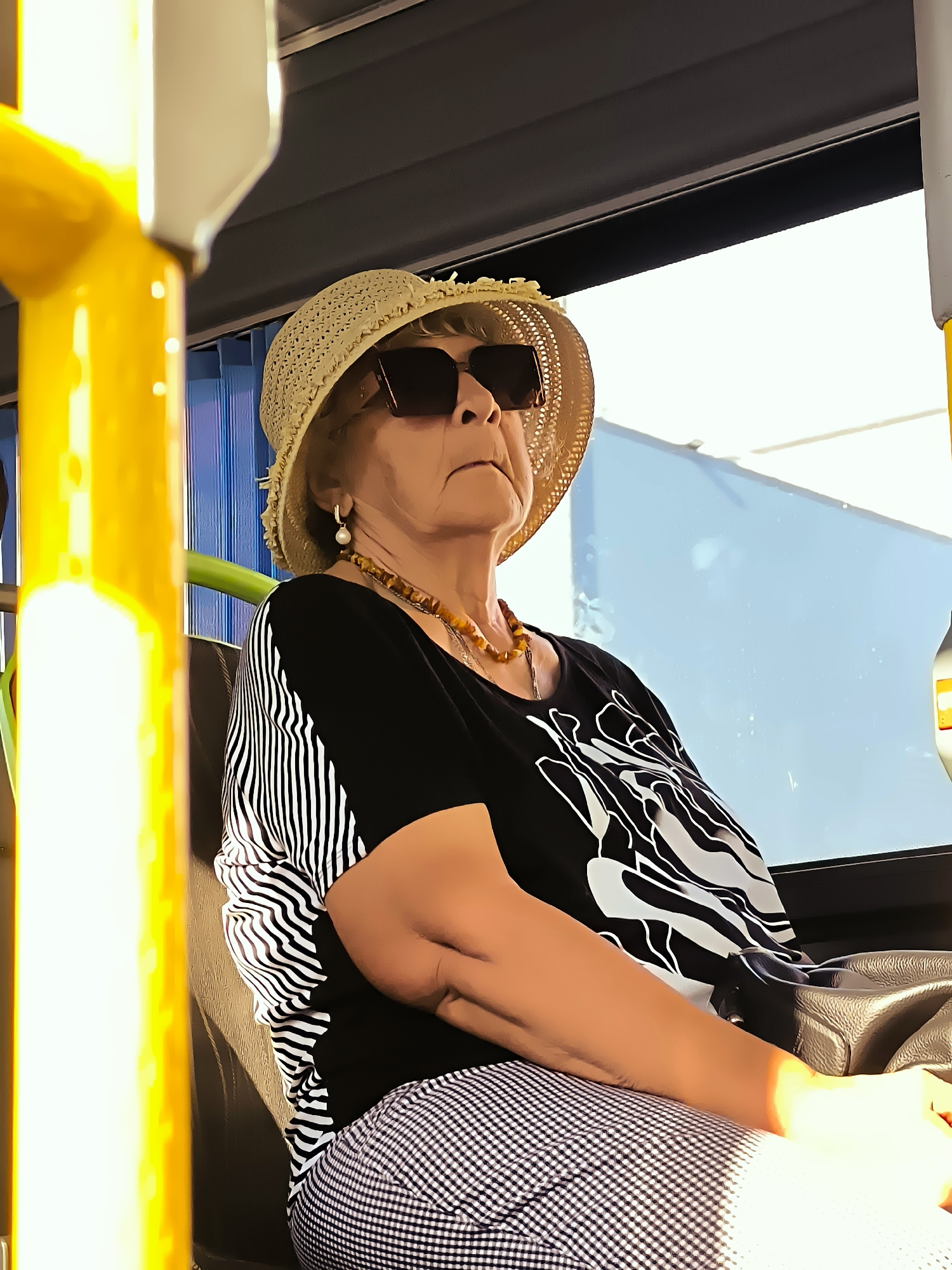 An elderly Caucasian woman wearing a straw hat and sunglasses is seated inside a bus. She has fair skin and is wearing a black t-shirt with white patterns and black and white checkered pants. She is also wearing a beaded necklace and earrings. The background shows the interior of the bus with a window and some yellow poles. The lighting suggests it is daytime and sunny.