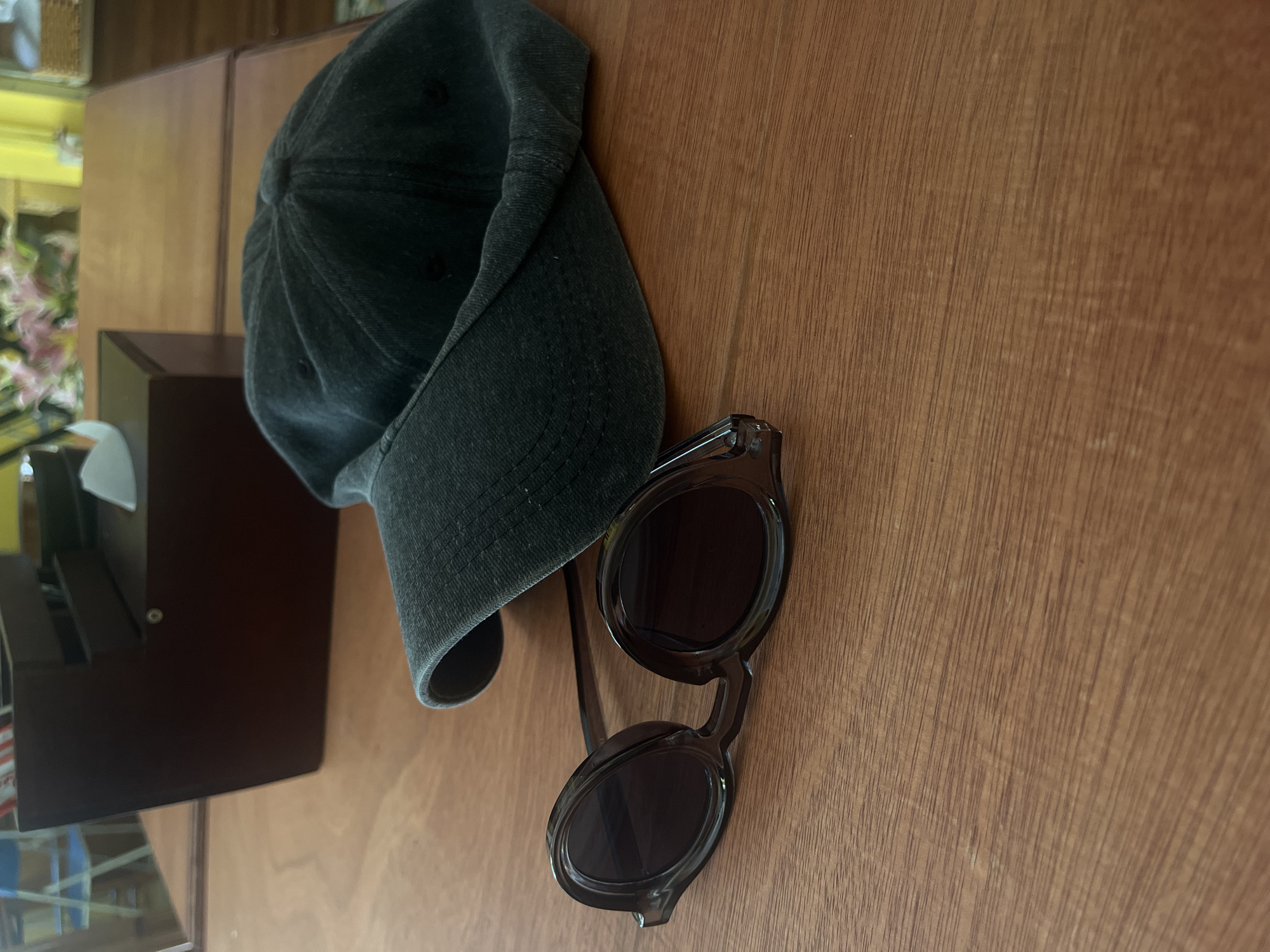 The image shows a dark green baseball cap and a pair of dark brown oval sunglasses lying on a wooden table. In the background, a dark wooden box with tissues and some blurry pink flowers are visible. The scene appears to be indoors. There is no information to suggest this is in Nha Trang Ward, Vietnam.