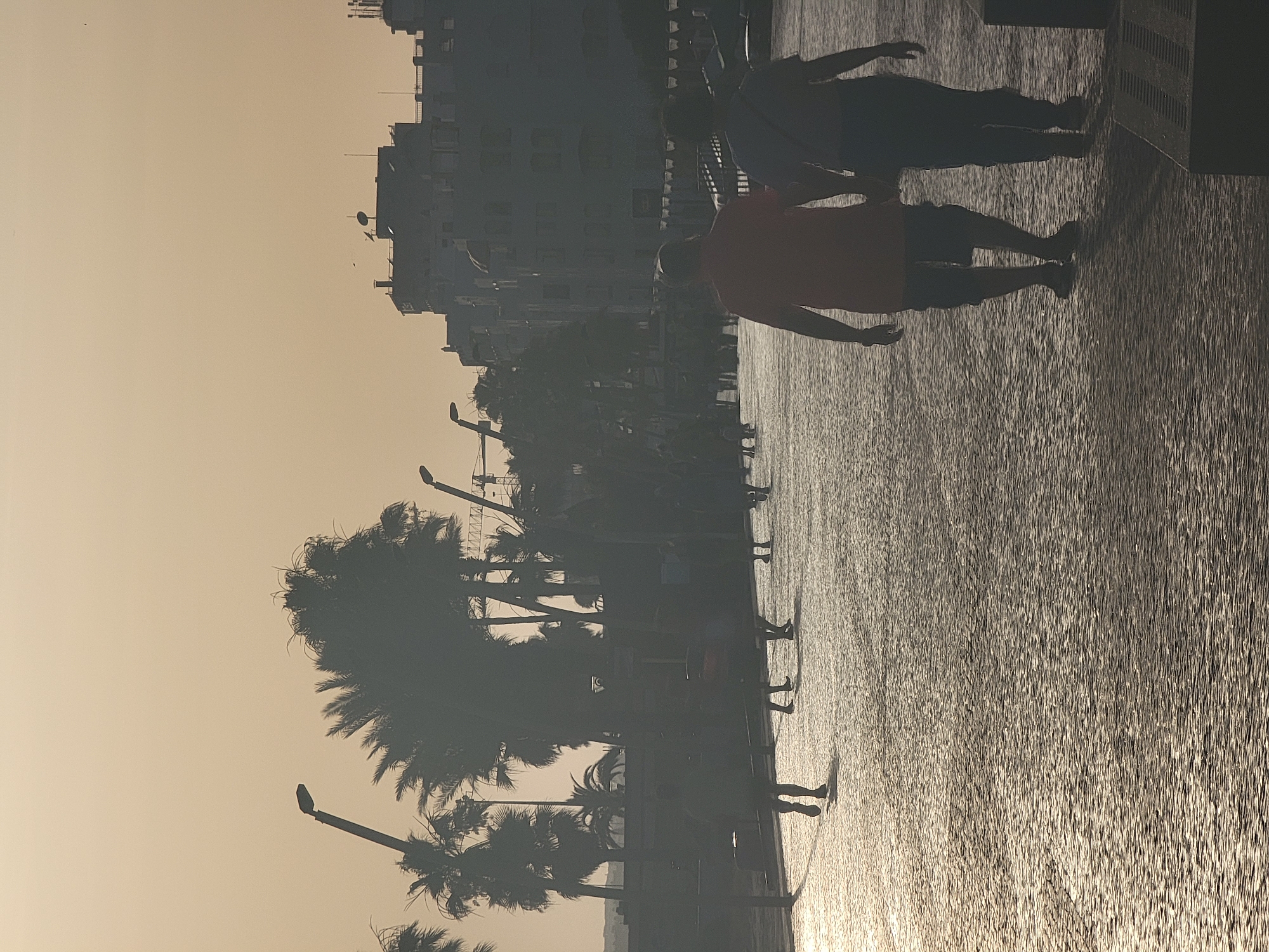 In Quarteira, Portugal, two people walk hand-in-hand on a cobblestone promenade as the sun sets. The sky is a hazy, warm orange, casting a soft glow on the scene. Palm trees line the promenade, their silhouettes dark against the sky. In the background, buildings and a construction crane are visible. The individuals are walking away from the camera, their figures silhouetted by the setting sun. The promenade is mostly empty, with a few other figures scattered in the distance.
