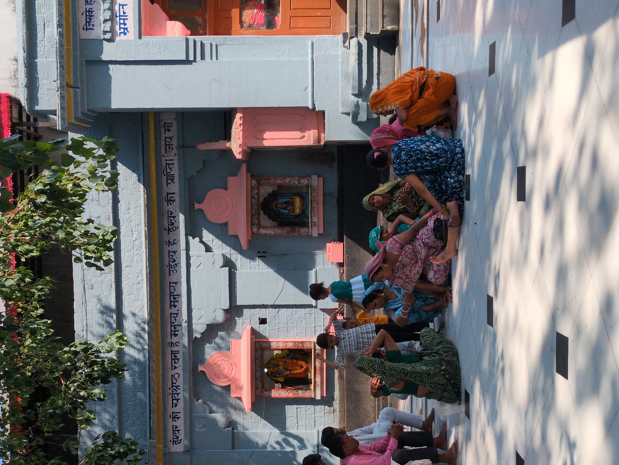 The image shows a street scene in the city of Kangra, India, with several people gathered in front of a building. The building appears to be a temple, with intricate carvings and decorative elements. The people are dressed in traditional Indian clothing, and they are interacting with each other in a relaxed and friendly manner. The scene is colorful and vibrant, with a sense of community and celebration. Some of the people are sitting on the ground, while others are standing. There is a small child in the center of the image, playing with a toy. The overall mood of the image is one of joy and happiness.