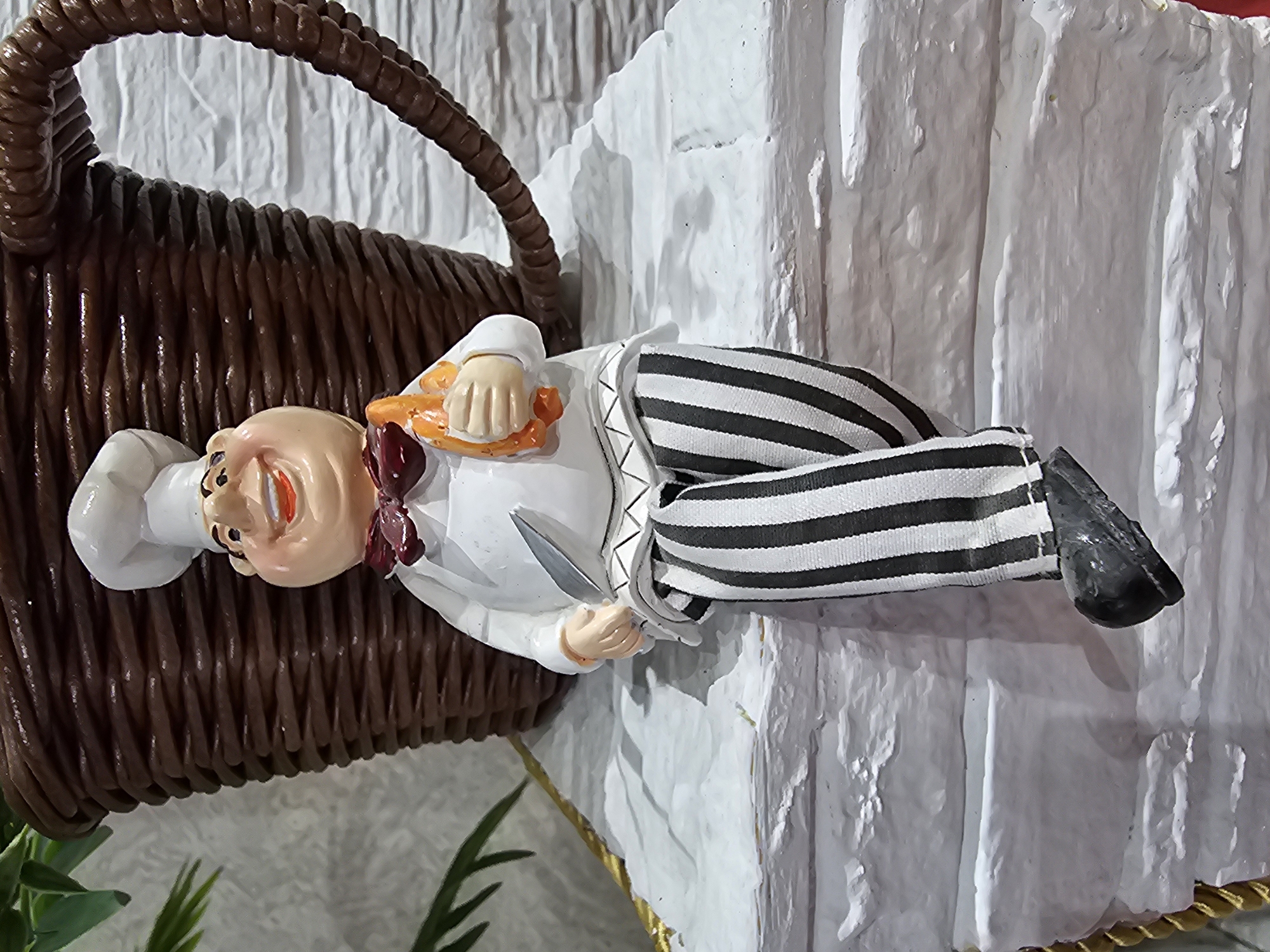 A ceramic figurine of a chef is perched on a white, textured surface resembling brickwork. The chef is wearing a white chef's hat, a white jacket with a dark red bow tie, and black and white striped pants. He is holding a knife in his right hand and a piece of orange food, possibly a pastry or bread, in his left hand. Behind the chef is a large, dark brown, woven-textured object, shaped like a coffee mug. The figurine is positioned in what appears to be an indoor setting, with a textured wall in the background and some greenery to the left. The scene is well-lit. There is no information available to suggest the location is the city of Kalinovka, Russia.