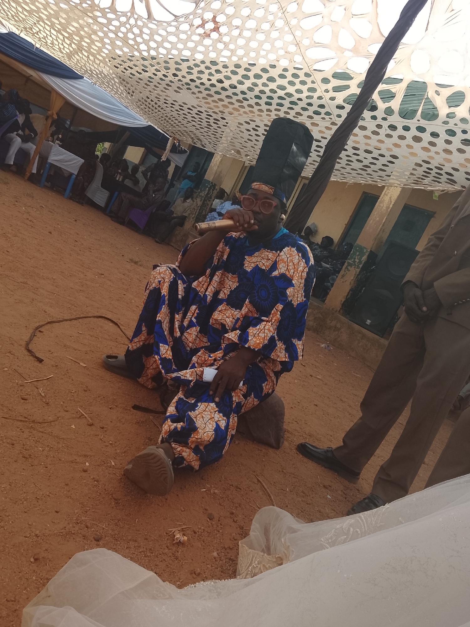 The image shows a man seated on the ground under a tent-like structure with a patterned covering. He is wearing a blue and orange patterned robe, red glasses, and a cap. He is holding a microphone to his mouth. A second person in a brown suit stands to his right. In the background, there are people sitting under additional tent structures. The ground is reddish-brown soil. A wedding dress lies in the foreground. Two black speakers are standing behind the man. The location is Unknown, Unknown.