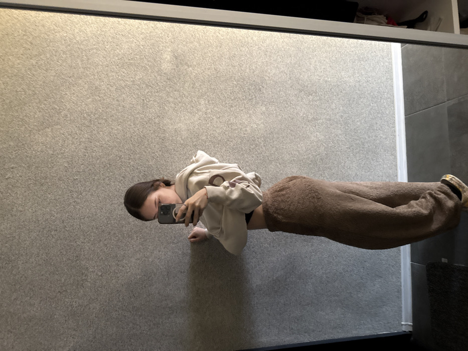 An individual is depicted taking a mirror selfie in an indoor environment. The subject, possessing dark hair, is visible from the upper chest to the feet, angled slightly to the left, with their face largely obscured by a dark-colored smartphone held in their right hand. Their left hand rests against a textured, light gray wall. The person is wearing a beige cropped hoodie, revealing a black undergarment and midriff, along with loose-fitting brown fuzzy pants. Yellow and black patterned footwear is partially visible on the right foot. The floor is dark gray, featuring a white baseboard where it meets the wall. A dark mat is present on the floor in the lower-left quadrant. A full-length mirror with a white frame reflects the scene, with a dark, indistinct area of the room visible beyond the mirror's edge on the right. The location within the city of Sofia, Bulgaria, cannot be determined from the provided visual evidence.