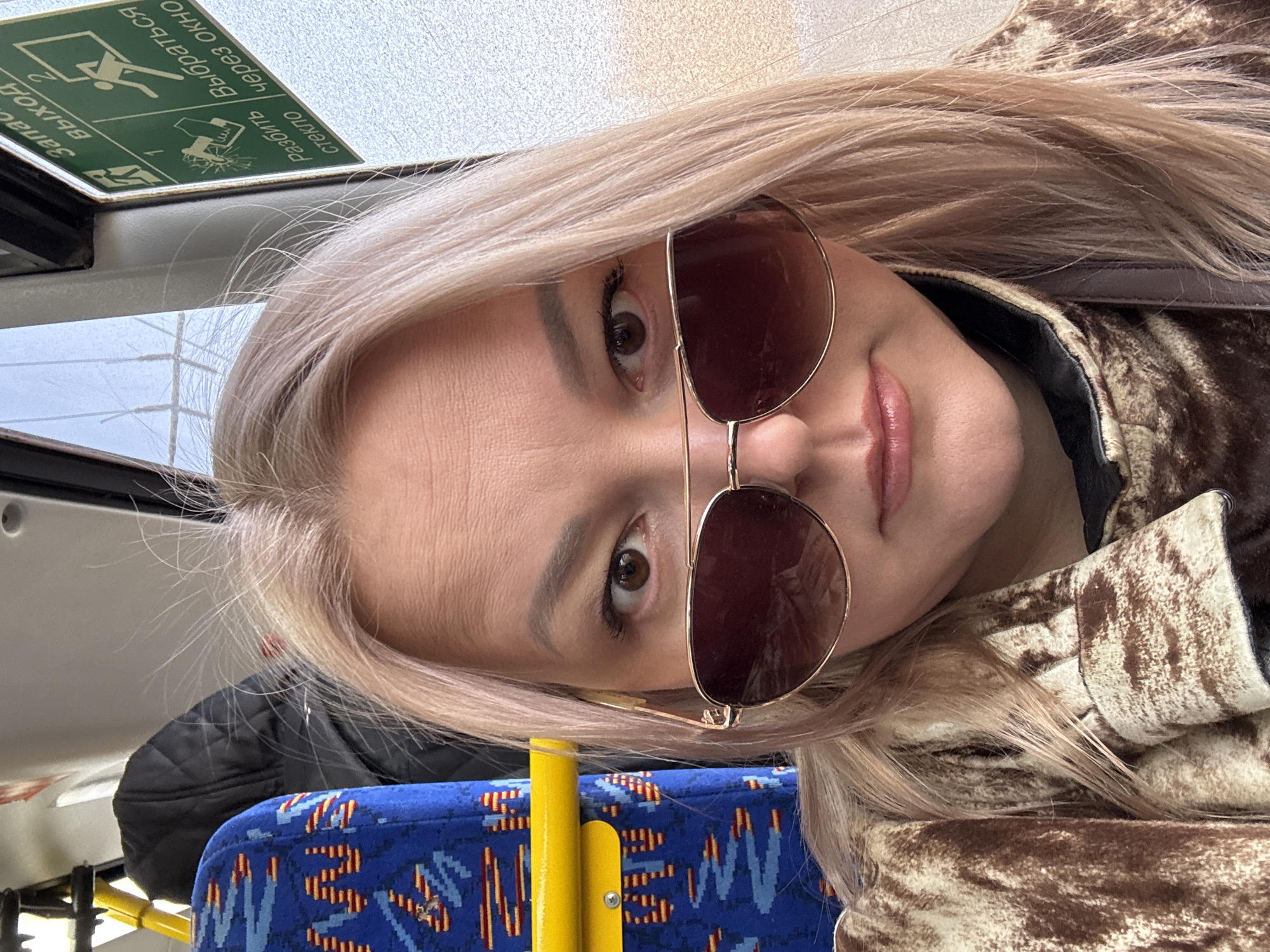 A woman with blonde hair, wearing sunglasses and a brown-and-beige patterned jacket, is seated on a bus in Izhevsk, Russia. The bus seat has a blue and yellow pattern. An emergency exit sign is visible above.