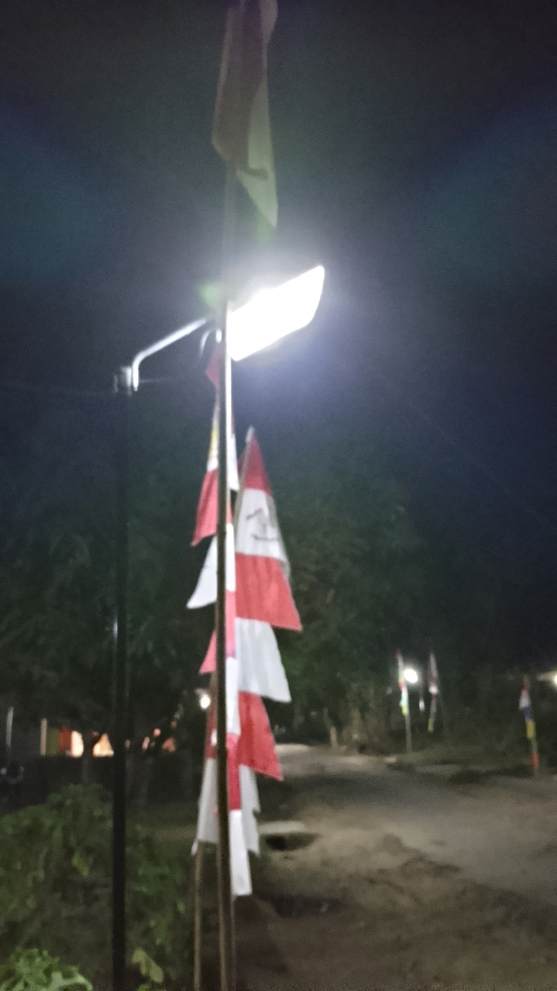 The image shows a dimly lit outdoor scene at night, with a bright lamppost illuminating a series of red and white flags hanging vertically from a pole. The flags appear to be Indonesian flags. The background consists of dark trees and a dirt path or road. Several other flags on poles are visible in the distance along the path, some of which are also illuminated by faint lights. The overall impression is of a celebratory or commemorative event taking place at night in a somewhat rural or park-like setting in Labuhan Ratu Delapan, Indonesia. There are no people clearly visible in the frame.