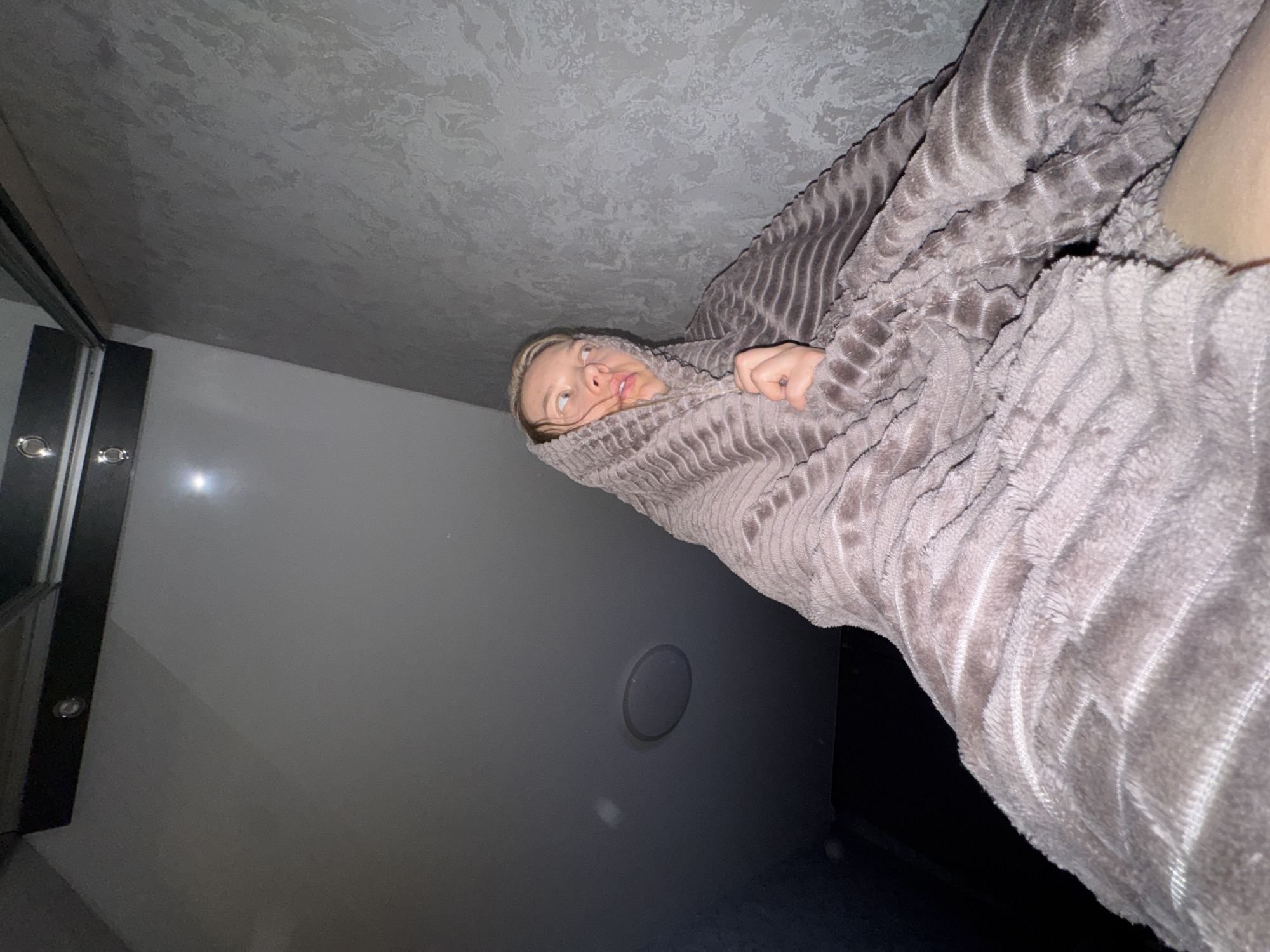 A person is wrapped in a textured, gray blanket, looking up and to the left of the frame. The person's face is visible, showing fair skin and light-colored hair. The background consists of a gray ceiling with a patterned texture, a light gray wall, and a dark-colored doorway or panel. A circular vent or access panel is visible on the light gray wall. The image appears to be taken from a low angle, looking upward. I don't have information about the location in the city of Chaikovsky, Russia.