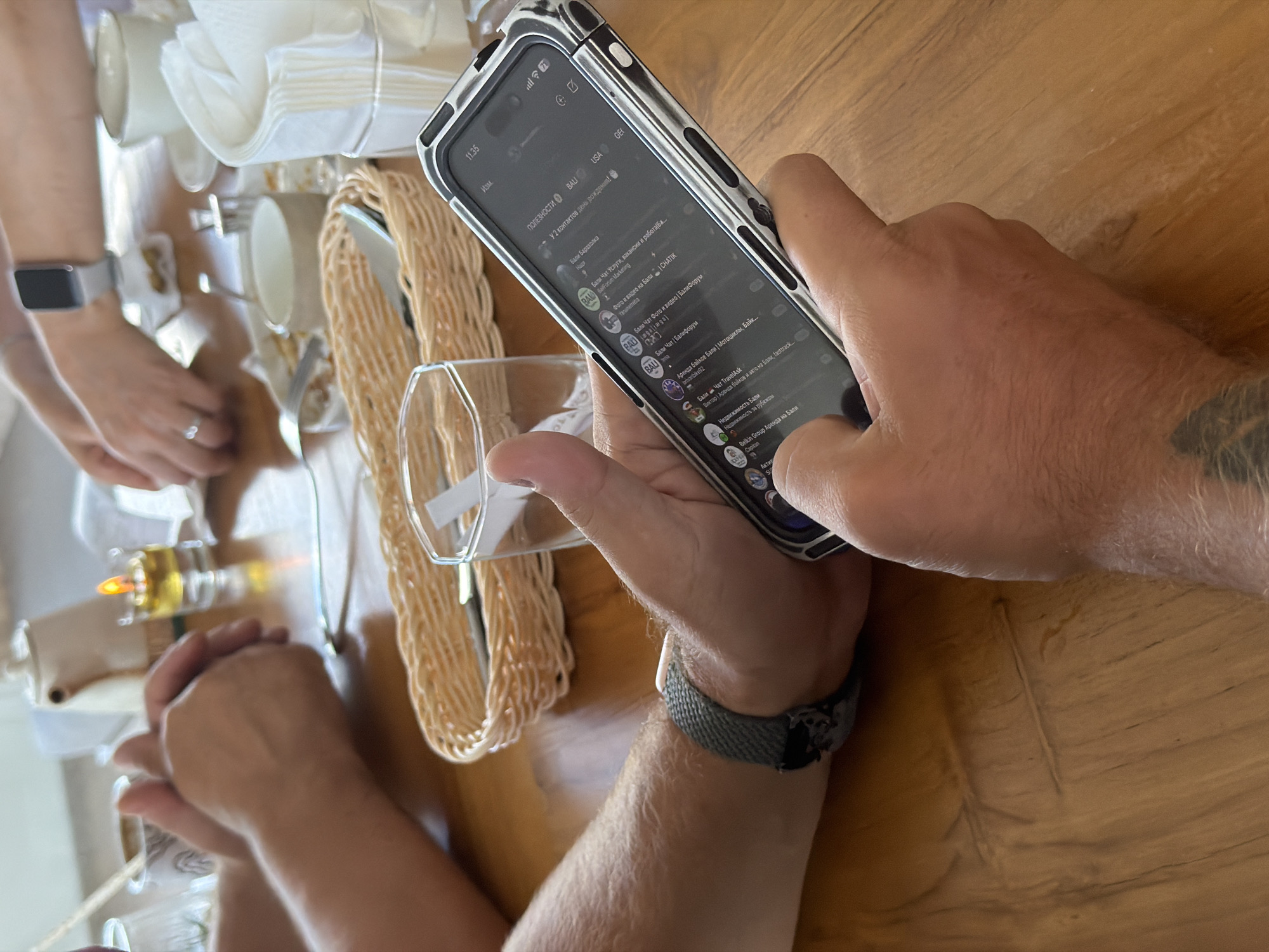 The image depicts a close-up view of a person's hands holding a smartphone, resting on a wooden table. The smartphone displays a messaging application with various chat conversations. In the background, other individuals' hands are visible, suggesting a social gathering. A small candle in a glass holder is lit on the table. Other items on the table include a woven basket containing cutlery, glasses, and napkins, indicating a meal or a break in an outdoor or semi-outdoor setting, possibly a cafe or restaurant. The overall lighting suggests it's daytime. The location is specified as Baturiti, Indonesia, though this detail is not visually verifiable from the image itself.