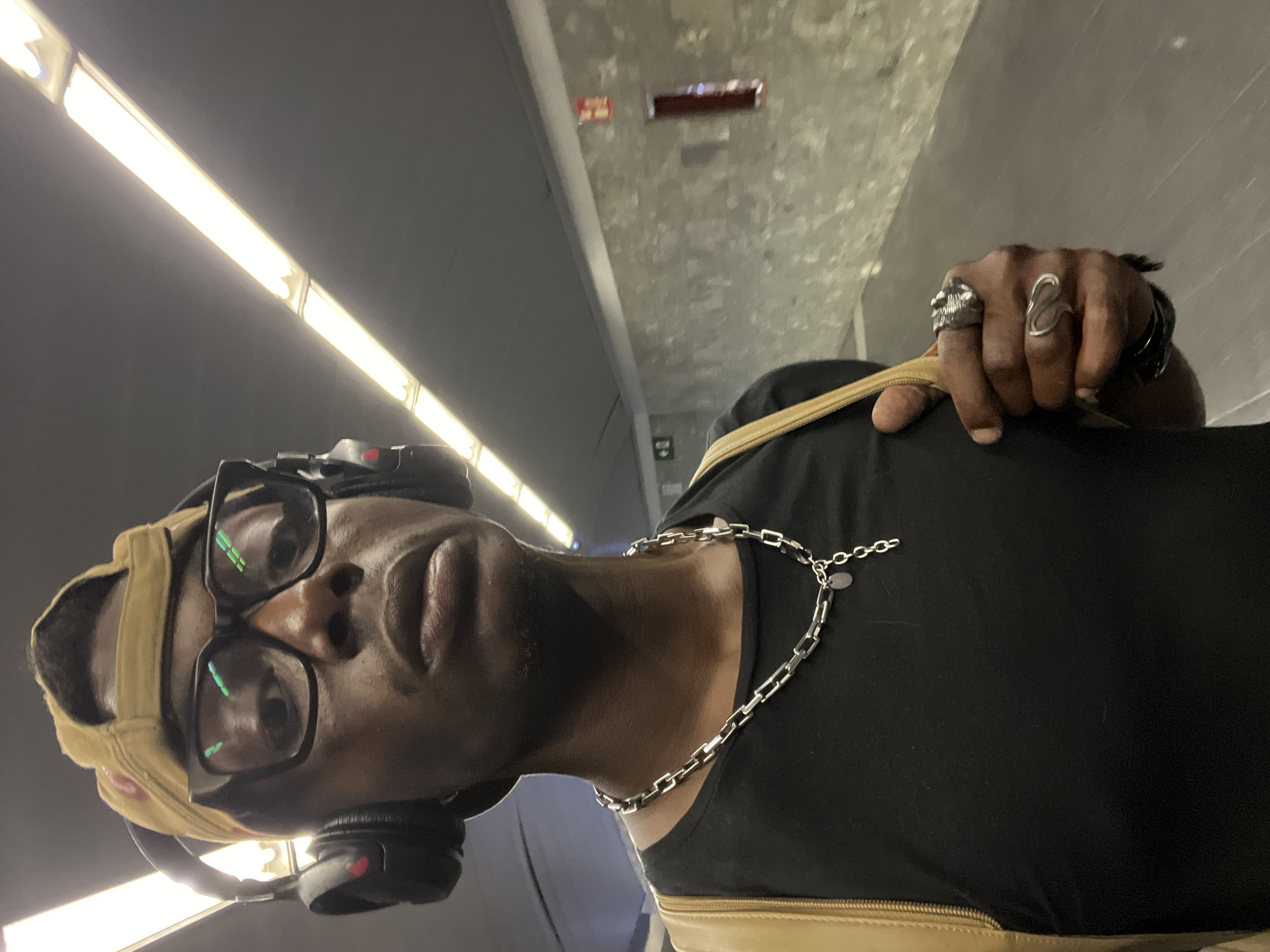 The central subject is a person with dark skin, captured in a low-angle selfie. They wear black-rimmed glasses, reflecting green and white light, black over-ear headphones, and a yellow baseball cap worn backward. A silver chain necklace with rectangular links and a small circular tag adorns their neck. On their right hand, a prominent silver skull ring and a distinct wavy silver ring are visible, as the hand rests on a light tan strap, likely from a bag.
The setting is an indoor space, characterized by a grey concrete ceiling and what appears to be a grey wall or upper structure. Long, bright fluorescent light fixtures illuminate the scene from above, running parallel to the line of sight. A small, reddish-brown rectangular vent or sign is visible high on the wall/ceiling. The environment suggests a modern, utilitarian interior, possibly a tunnel, corridor, or station.
The person is looking directly at the camera, indicating they are taking a selfie or posing for a photograph. The time of day is not discernible due to the artificial indoor lighting, and weather conditions are not visible. The person's expression is neutral to serious. No clear, readable text is visible in the image.