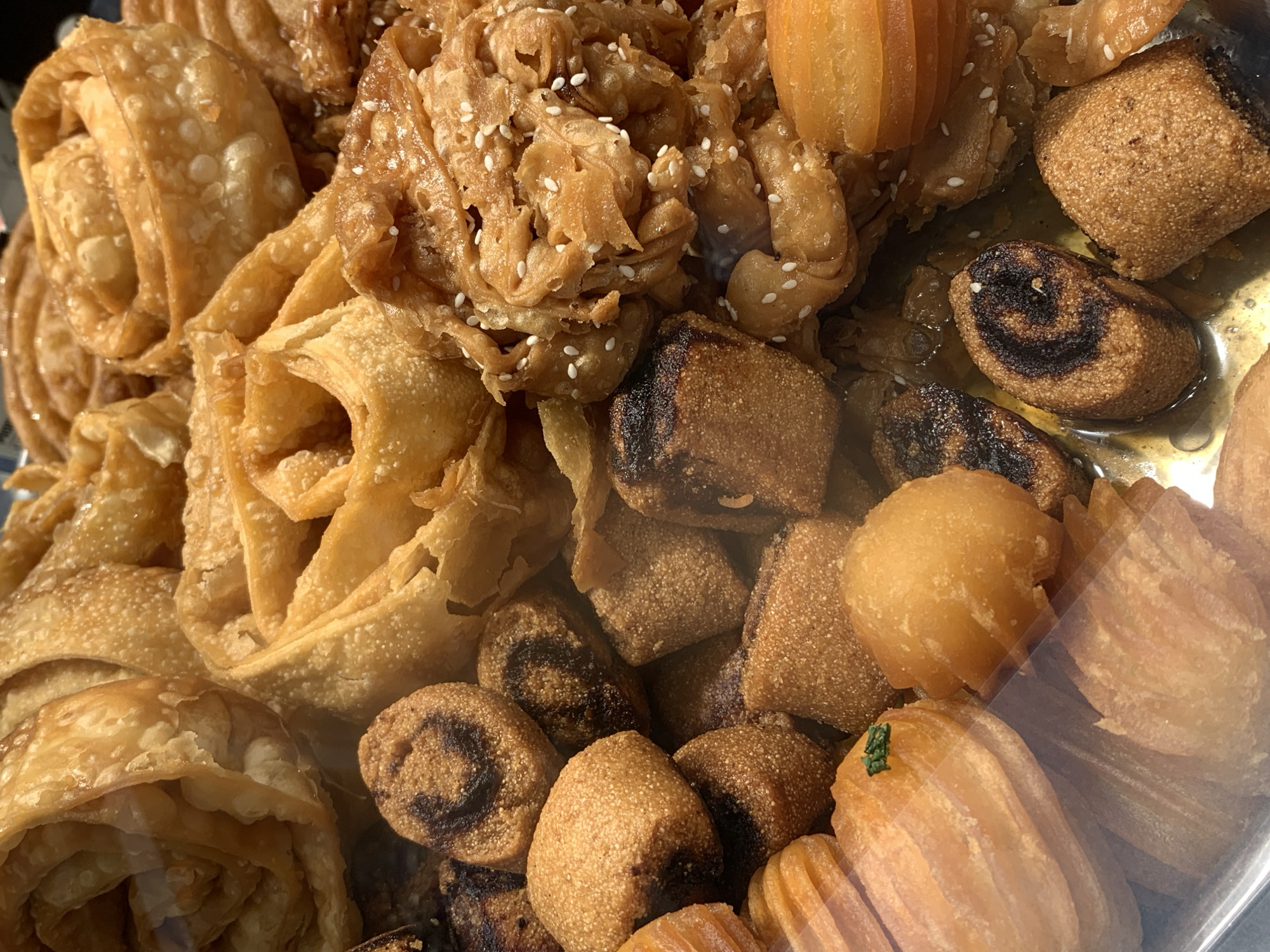 This is a close-up shot of a selection of Middle Eastern pastries. There are several varieties, including what appears to be a type of rose-shaped fried dough, topped with sesame seeds, cube shaped, and a type that resembles small, elongated cylinders. All the pastries appear to be glistening, likely due to a sweet syrup. The image was taken in Tel-Aviv, Israel.