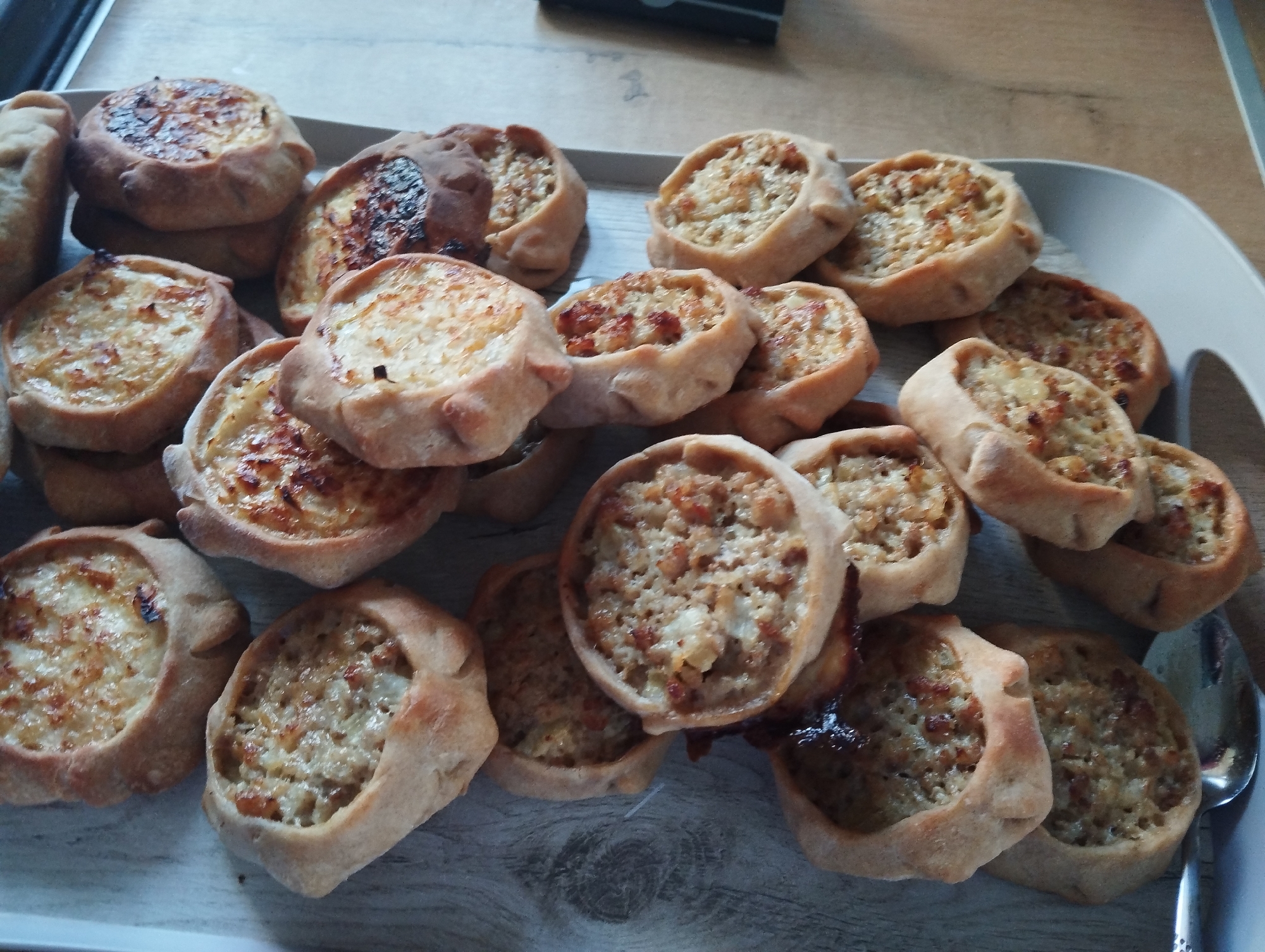 A tray of Karelian pies (karjalanpiirakka) is on a light-colored wood-grain surface. The pies vary in filling color, from light to dark. One pie is severely burnt. A stainless steel spoon is in the lower right corner. The location is likely a home or restaurant kitchen, but it is not possible to verify the specific location in Krasnogorskoye, Russia. There are no people visible in the image.