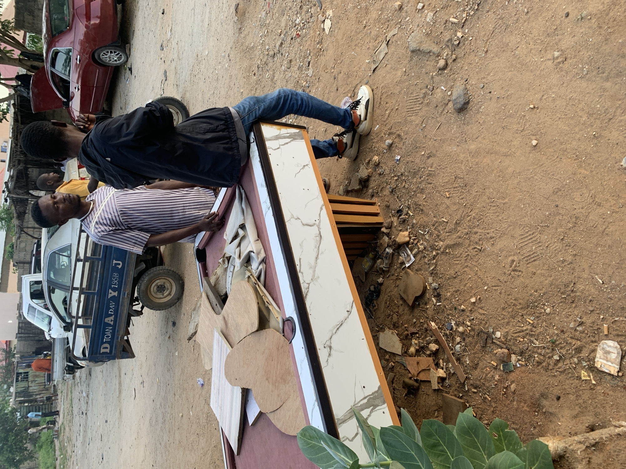 The scene depicts an outdoor area in Adankolo, Nigeria, with a dirt ground. A dismantled pool table is the central object, along with various pieces of wood and discarded materials scattered around it. Two men stand near the table; one wears a striped shirt, and the other a black jacket and jeans. A pickup truck is visible in the background. A red car is partially visible in the background.
