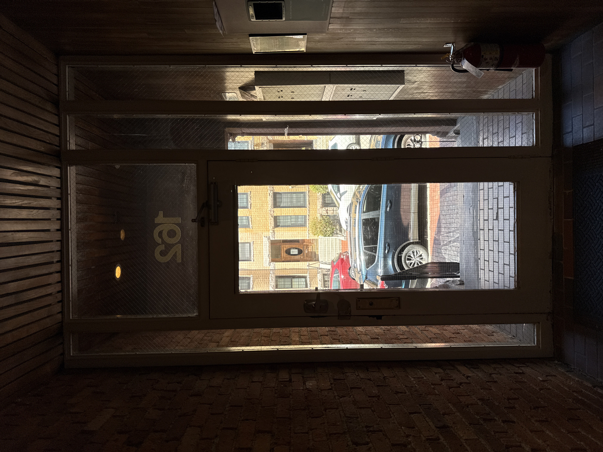 The image shows a view from inside a building, looking out through a glass door/window at an urban scene. A building facade is visible across the street. Cars are parked on the street, including a dark colored minivan and a red vehicle. A fire extinguisher is mounted on the ceiling inside the building. The door has the letters "ies" on it. Based on the presence of "ies" on the door, this appears to be a view from the Boston University Institute for Economic Studies building in Boston, United States of America.