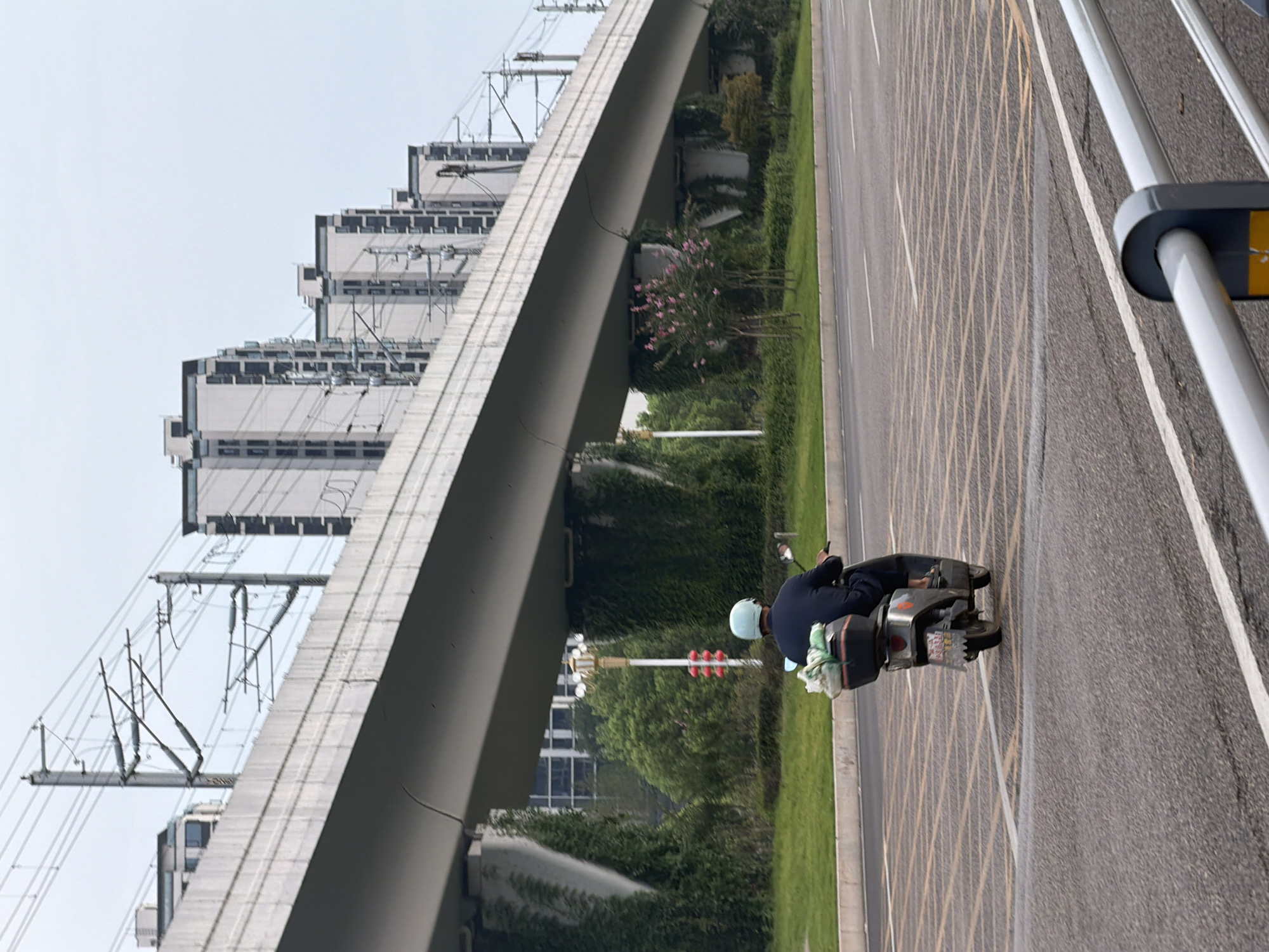 A person wearing a blue helmet rides a scooter down a road in Jiangjin, China. The scooter has a black storage box on the back, which is carrying a bundle of items wrapped in green plastic. The road has yellow grid markings. In the background, a raised highway or train track structure is supported by concrete pillars covered in ivy. Beyond the elevated structure, a row of modern, multi-story buildings with dark-trimmed windows are visible. The sky is overcast.