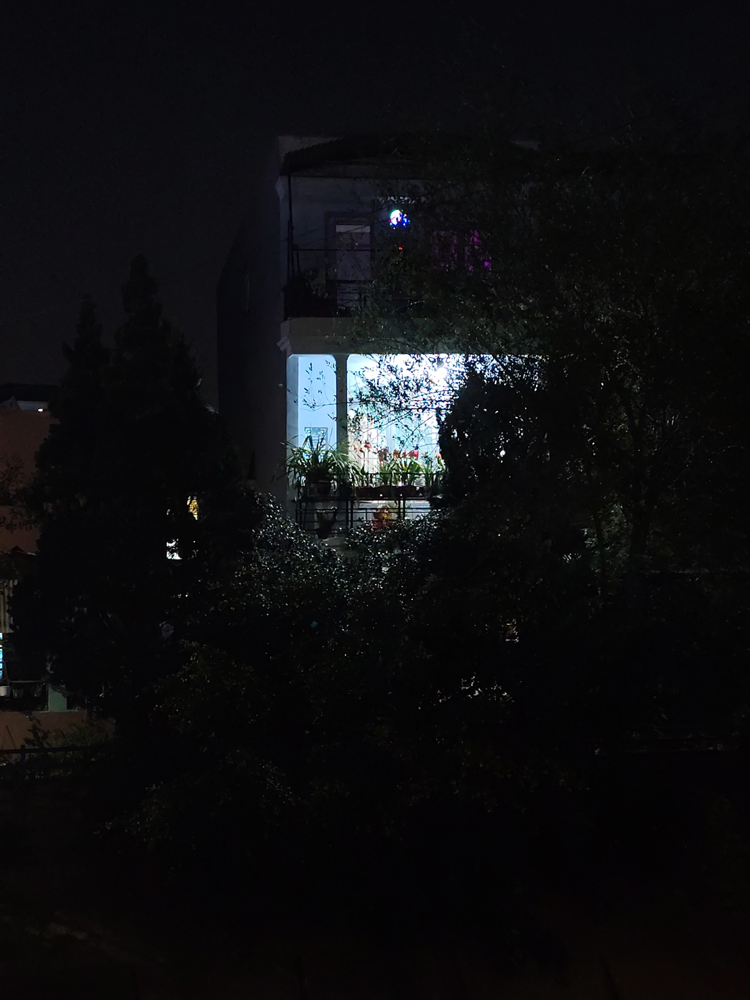 At night in Da Lat, Vietnam, the image captures a brightly lit balcony of a multi-story building framed by dark trees. The balcony contains several potted plants and appears to be decorated. Above the lit balcony, smaller lights are visible on an upper balcony. The foreground consists primarily of dark vegetation, obscuring the lower portion of the building. The overall scene is dark, with the light emanating from the balcony drawing the eye. The presence of other buildings and city lights is minimal.