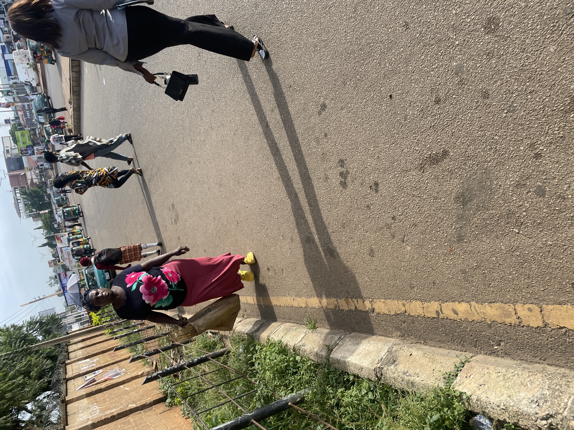 The image shows a street scene in Jos, Nigeria. In the foreground, a woman wearing a dark blue t-shirt with large pink floral patterns and a long pink skirt walks on the right side of the road, carrying a large bag of what appears to be grass or reeds over her left shoulder. She is wearing yellow shoes. To her right, another woman in a grey blazer and black pants walks away from the camera, carrying a black handbag.
In the background, other people are walking and various vehicles, including what appear to be tricycles and cars, are present on the street. Buildings and some vegetation line the side of the road. The sky is visible above, indicating it is daytime. A low concrete barrier with a yellow line separates the sidewalk from the road. The overall scene depicts a busy urban environment.