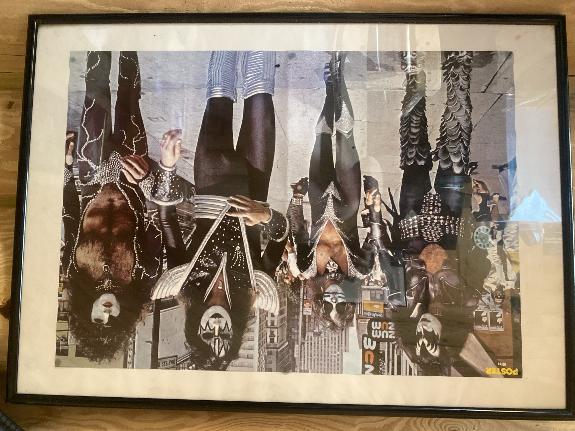 The image shows a framed poster of the rock band Kiss posing together in the middle of a city street. The band members are dressed in their signature elaborate costumes, featuring platform boots, studded outfits, and heavy makeup. They are walking down a sidewalk, with buildings and other pedestrians visible in the background. The poster has a yellow "POSTER" label in the upper left corner, with "Kiss" written below it. There are no visible elements that associate this image with Lindo-Pustyn' (Kantaurovskii S/S), Russia.