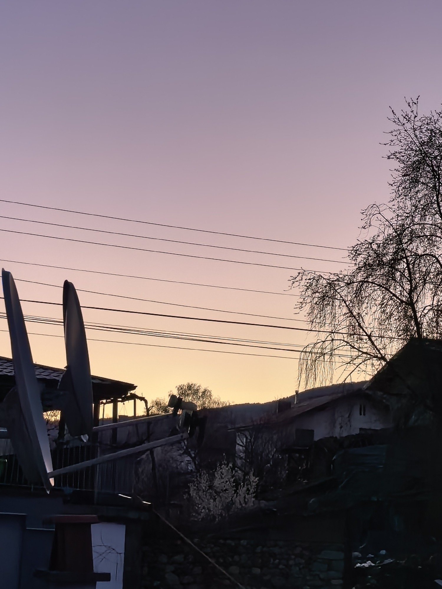 The image depicts an outdoor scene at dusk. The sky is a gradient of pink and orange, with power lines stretching across it. Two satellite dishes are visible in the foreground, positioned on a roof or structure. Below the horizon line, there are silhouetted structures resembling houses or buildings, along with trees and vegetation. The scene appears to be in a residential area.