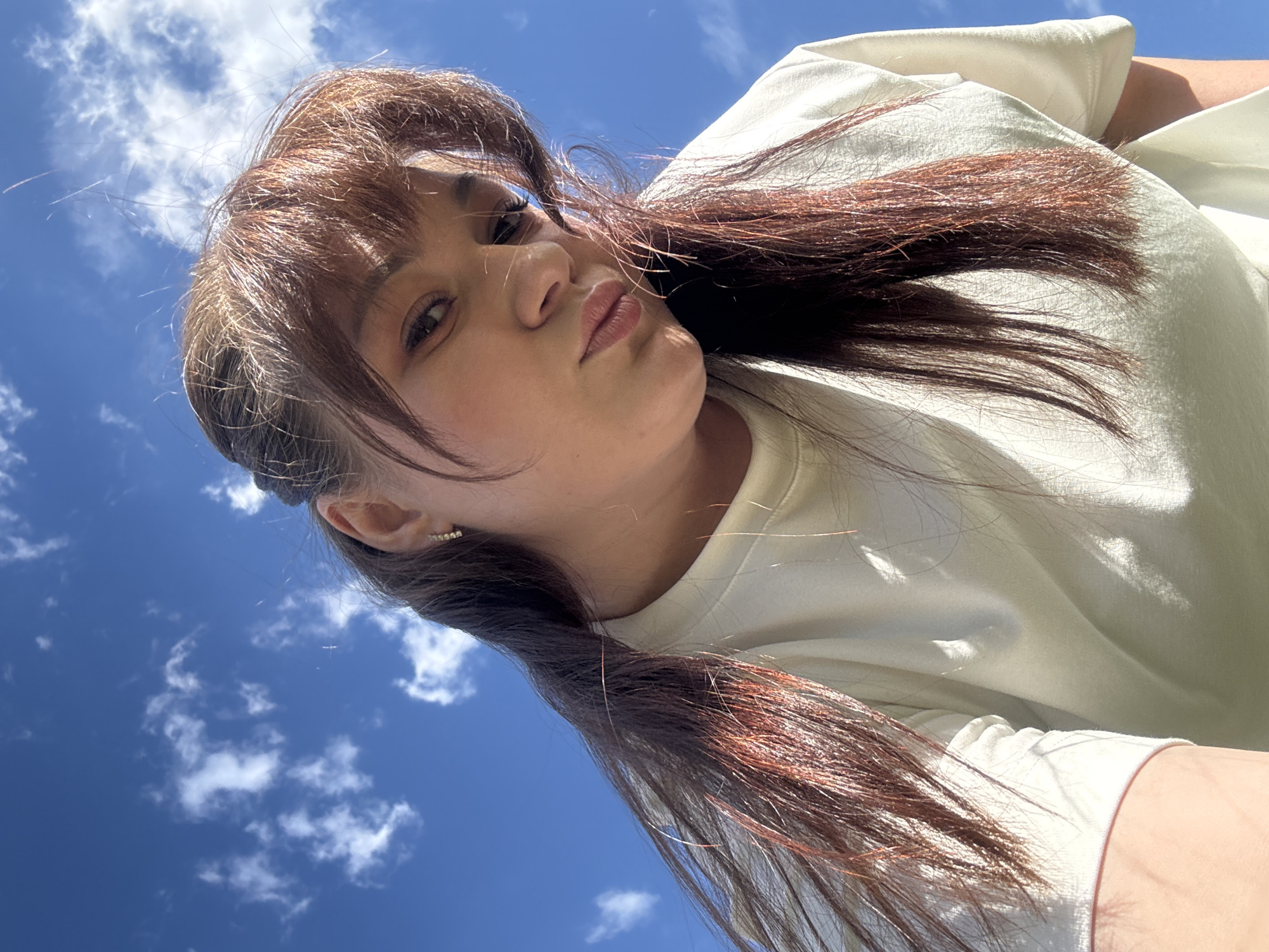A young woman is captured in a low-angle, wide shot outdoors on a sunny day. She is wearing a light-colored t-shirt and her long, reddish-brown hair is spread out around her. Her face is turned towards the camera, with her eyes looking forward and her lips pursed. She is wearing a small, sparkling earring on her visible ear. The background is dominated by a bright blue sky with scattered white clouds, suggesting good weather. The angle of the shot makes it appear as though she is looking up at the sky. There are no visible text elements in the image. The lighting suggests it is daytime, likely mid-morning or afternoon, given the brightness of the sky and the shadows cast on her clothing. The scene conveys a casual and perhaps playful mood.