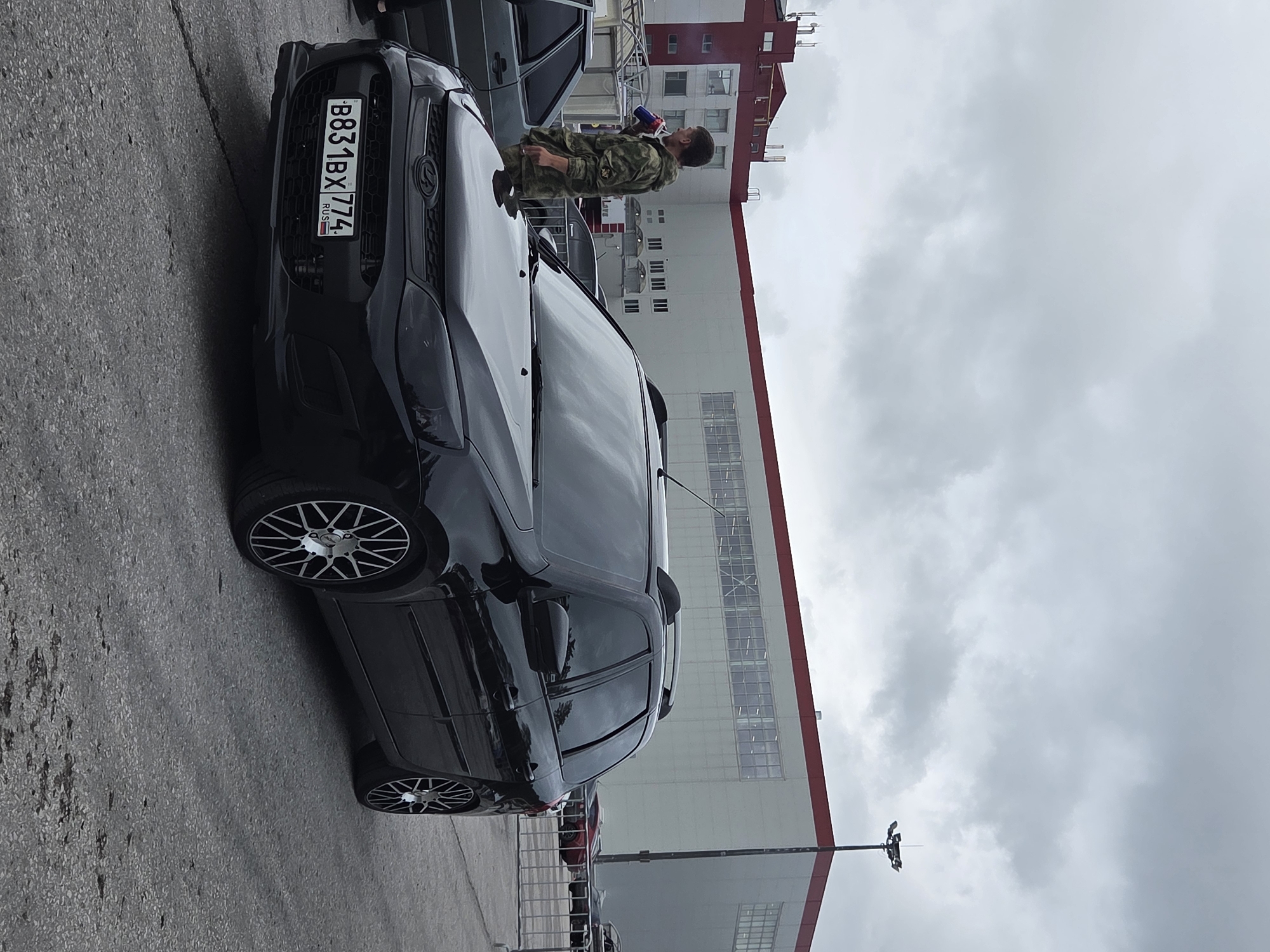 This image captures an outdoor scene, likely a paved parking area, beneath an overcast sky. In the foreground, a black Lada Kalina wagon is prominently displayed. The vehicle features modifications including blacked-out headlights, tinted windows, roof rails, and aftermarket multi-spoke alloy wheels. Its front license plate, "B831BX 774 RUS", indicates registration in Chelyabinsk Oblast, Russia.
To the left of the black Lada, a male individual is visible, dressed in camouflage fatigues. He is holding a large blue and red container, possibly a beverage cup, near his mouth. A portion of a grey older-model Lada is visible behind him on the far left.
In the mid-ground, a metal barrier runs horizontally, separating the immediate foreground from a background area. Beyond this barrier, several other vehicles are partially discernible, including a red car. The background is dominated by a large, multi-story building with a light-colored facade and distinctive red trim along its roofline and some lower sections. The building has numerous windows, some appearing as large industrial panes. A tall light pole, equipped with multiple floodlights, stands to the far right, adjacent to the building. The ground is asphalt, showing visible cracks and dark, irregular stains. No specific visual information within the image definitively identifies the location as Perm, Russia.
