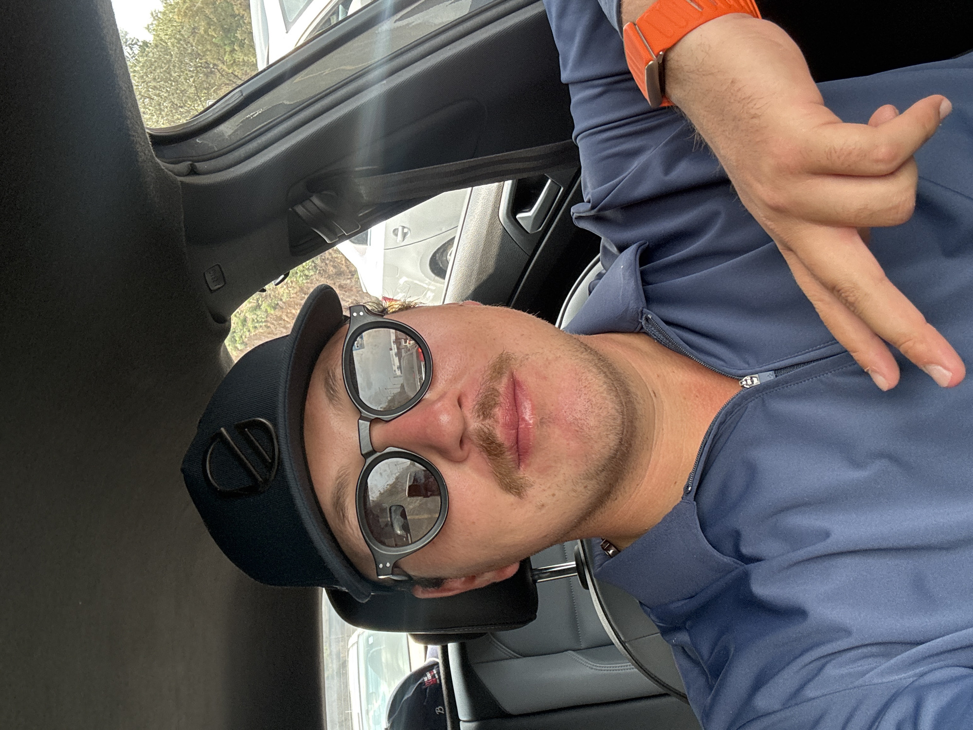 A man wearing a navy blue cap with a logo and round sunglasses sits in a car. He is wearing a navy blue jacket and an orange smartwatch. He is holding his hand up with two fingers extended. The interior of the car is visible. The man is likely located in Huixquilucan de Degollado, Mexico, but the image does not contain enough information to confirm this.