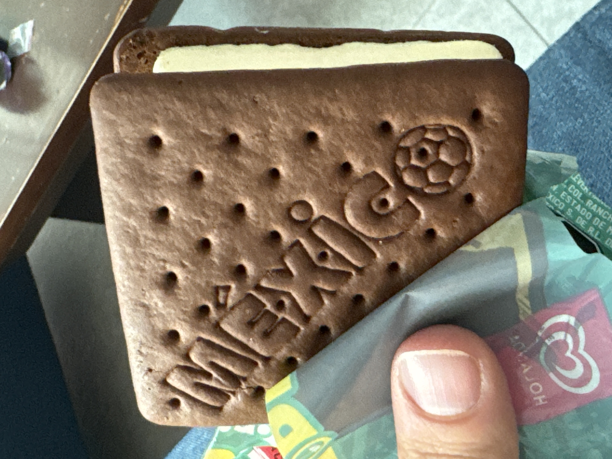 A close-up photograph shows a chocolate-colored ice cream sandwich with the word "México" and a soccer ball embossed on the top cookie. The ice cream sandwich is being held by a finger, partially unwrapped from its packaging, which has a pink logo on a green and blue background. In the background is a table and a leg with a jeans. The text on the packaging indicates that the ice cream was made in San Bernardino Tlaxcalancingo, Mexico.