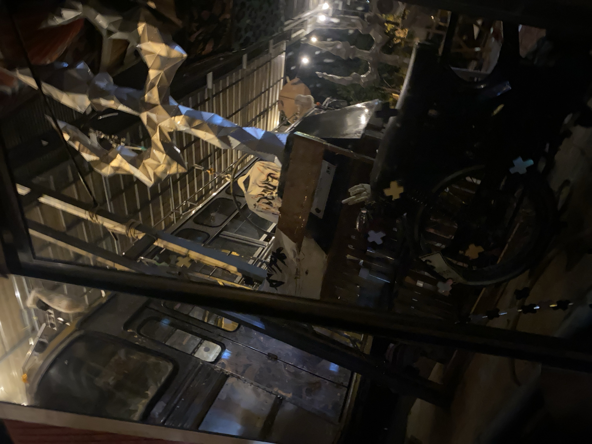 Captured from a low-angle perspective, the photograph depicts a dark and cluttered outdoor scene. The primary object in view is a vintage bus with a weathered, metallic exterior. The bus appears to be stationary and possibly repurposed as part of a decorative or artistic installation. The environment includes various metallic and sculptural elements, creating an industrial or avant-garde aesthetic. The image is taken at Baan Nai Suan, Thailand.