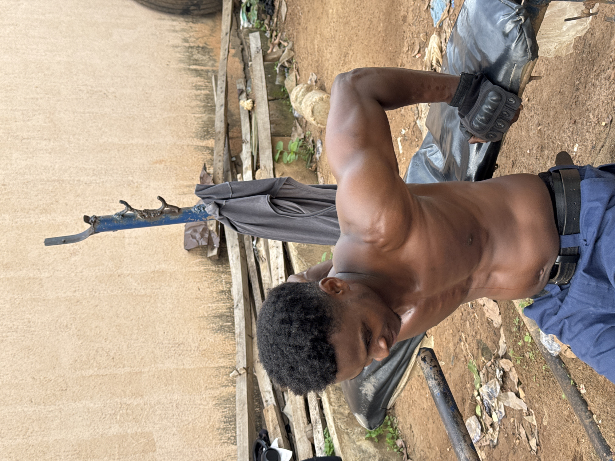 A shirtless Black man with dark curly hair is performing a dip exercise on a black vinyl workout bench outdoors. He is wearing blue pants and black fingerless gloves. The man is leaning forward with his elbows bent, supporting his weight on his hands. The background shows a textured, beige wall, stacks of wooden planks, and some debris on the ground. The scene appears to be in a simple, outdoor gym environment, possibly in Akwanga, Nigeria, given the context provided.