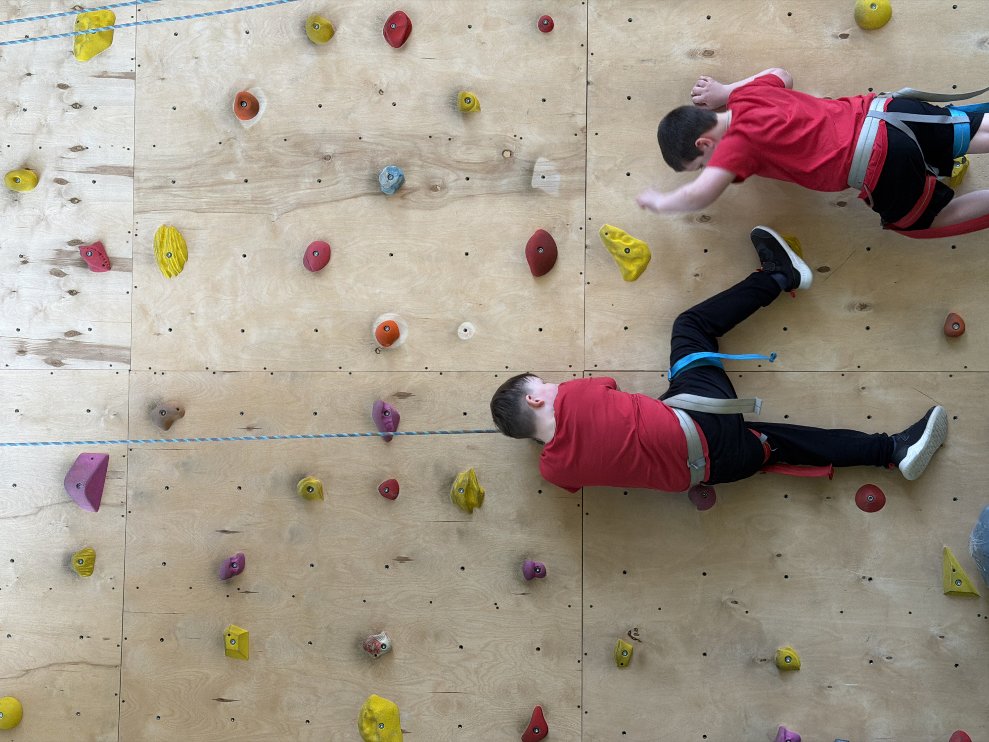 Two boys are climbing an indoor climbing wall. The wall is constructed of light-colored wood panels with colorful climbing holds attached. Both boys wear red shirts, harnesses, and athletic shoes. One boy is positioned higher on the wall, reaching with his right hand. The other boy is lower, with his left hand on a climbing hold and legs extended. A blue and white rope is visible horizontally across the wall. The location is Izhevsk, Russia.