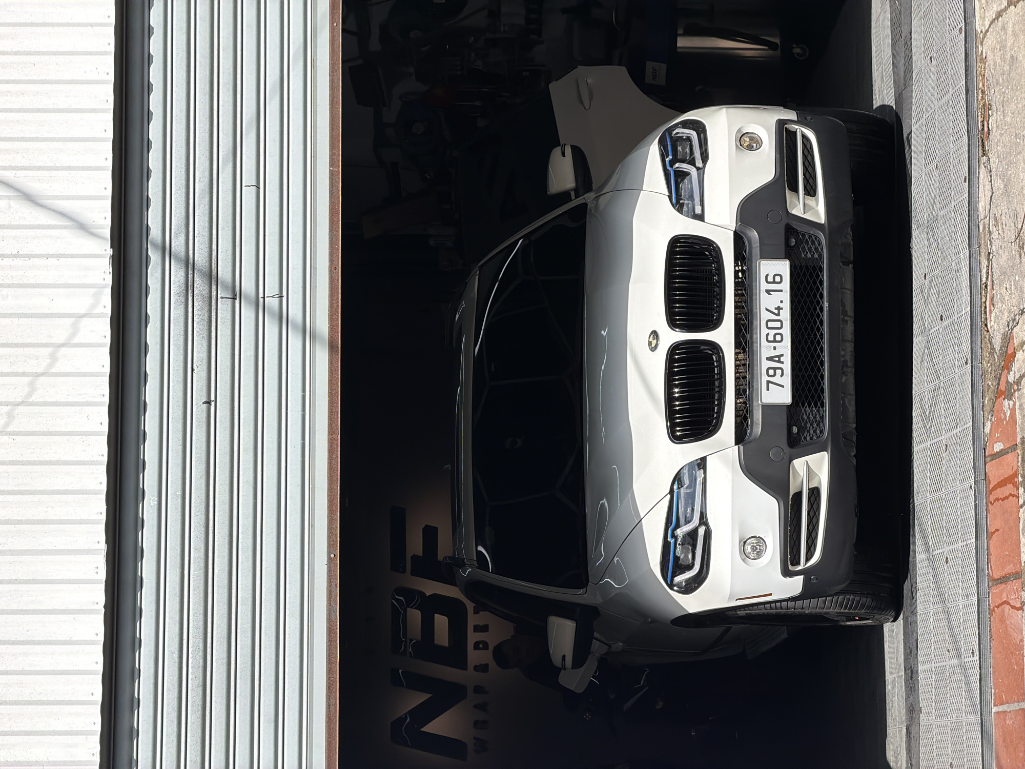 A white BMW X5 SUV is parked inside a garage with a partially open roll-up door. The vehicle's front is prominently displayed, featuring its grille, headlights, and license plate which reads "79A-604.16". The garage interior appears to be a workshop, with equipment and tools visible in the background. A person is partially visible on the left side of the vehicle, leaning towards it. The floor is a patterned grey surface, with reddish brickwork visible in the foreground. The overhead lighting casts shadows, and the overall scene suggests a commercial automotive service location. The location is identified as NBF Wrap & Detail in Bac Nha Trang Ward, Vietnam.