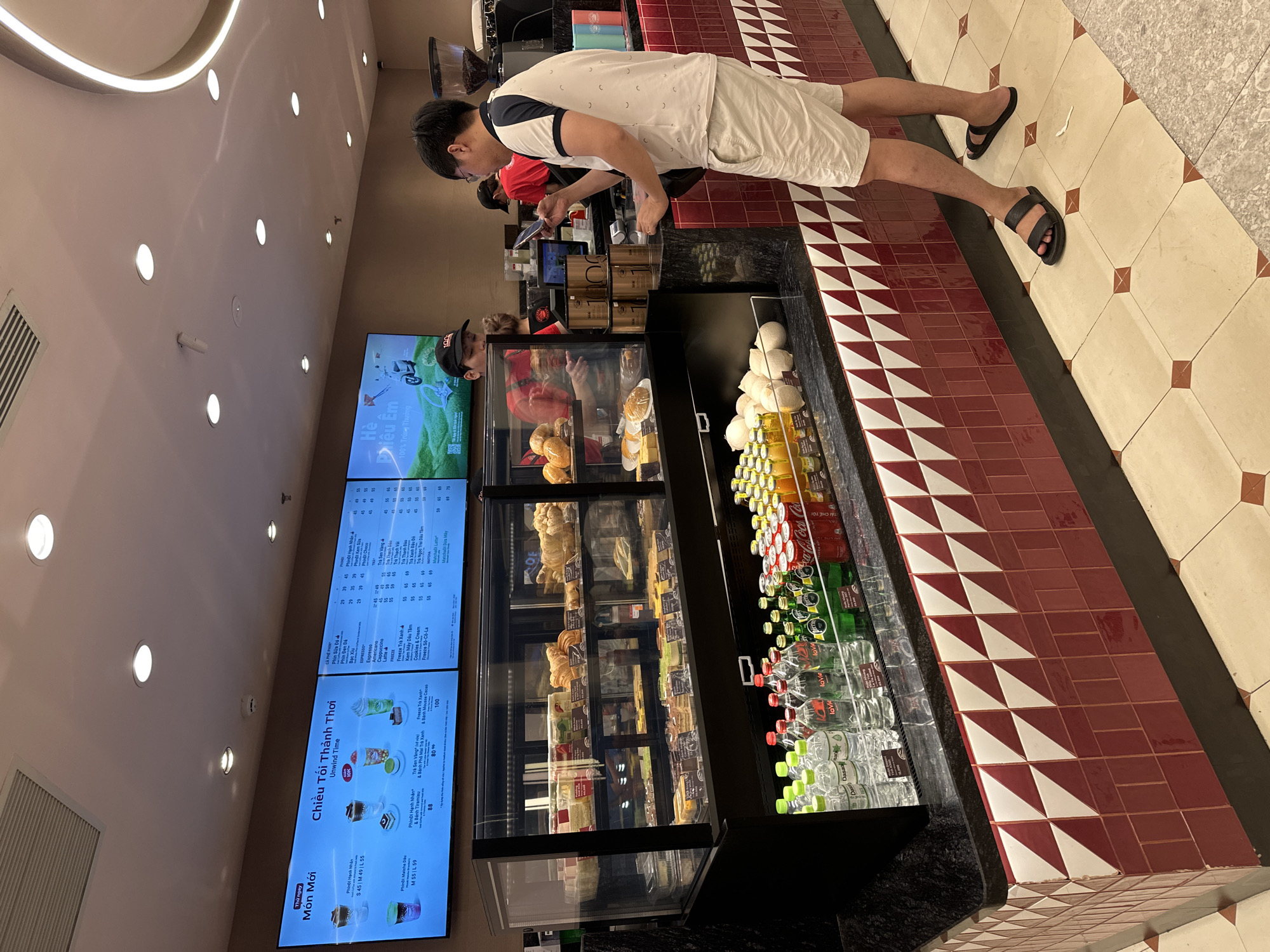 This is a vertical shot of the interior of a cafe, likely in Da Nang, Vietnam, based on the Vietnamese text. The scene is brightly lit, suggesting it's daytime or the cafe is well-illuminated.
In the foreground, a man wearing glasses, a white t-shirt with a subtle pattern, and light-colored shorts stands with his back to the camera, facing a counter. He is wearing black sandals. He appears to be looking at a menu or interacting with staff. Behind him, other individuals can be seen, including someone in a red shirt.
The counter area features a display case filled with pastries like croissants and cakes. To the right of the display case, a cooler is stocked with bottled drinks, including water and what appear to be soda bottles, some with green labels. Above the counter, large digital screens display menus with Vietnamese text and images of drinks and food. One screen prominently shows "Chiều Tối Thảnh Thơi" (Evening Relaxation) and drink specials.
The floor is tiled with a pattern of red and white triangles and squares. The overall atmosphere suggests a typical cafe setting where customers order drinks and snacks. The focus is on the ordering area, highlighting the food and drink offerings.