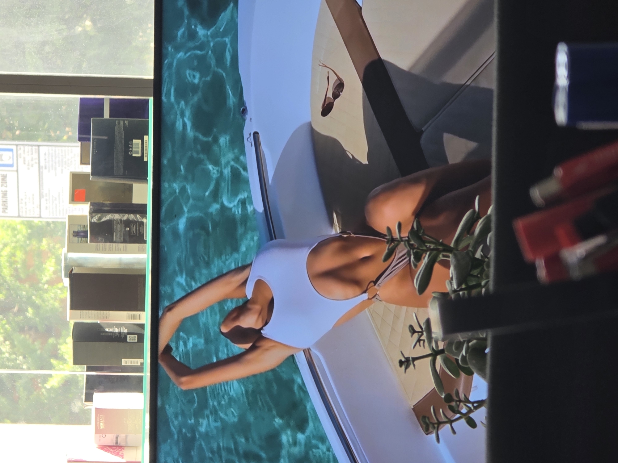 The image depicts a person in a white swimsuit reclining on a boat. The boat appears to be on clear, turquoise water, suggesting a sunny day, possibly at a tropical or Mediterranean location. A pair of sunglasses rests on the boat's surface. In the background, to the left of the boat, there is a display of various boxes, some of which appear to be perfume or cosmetic products. Text on one of these boxes is partially visible, showing "BVLGARI". Further left, a sign with the words "PARKING ZONE" is visible. On the right side of the image, a shelf contains several items, including what look like makeup or toiletries. The overall scene evokes a sense of leisure and luxury. The location context is Varna, Bulgaria, which can be a summer tourist destination. The time of day appears to be daytime, given the lighting.