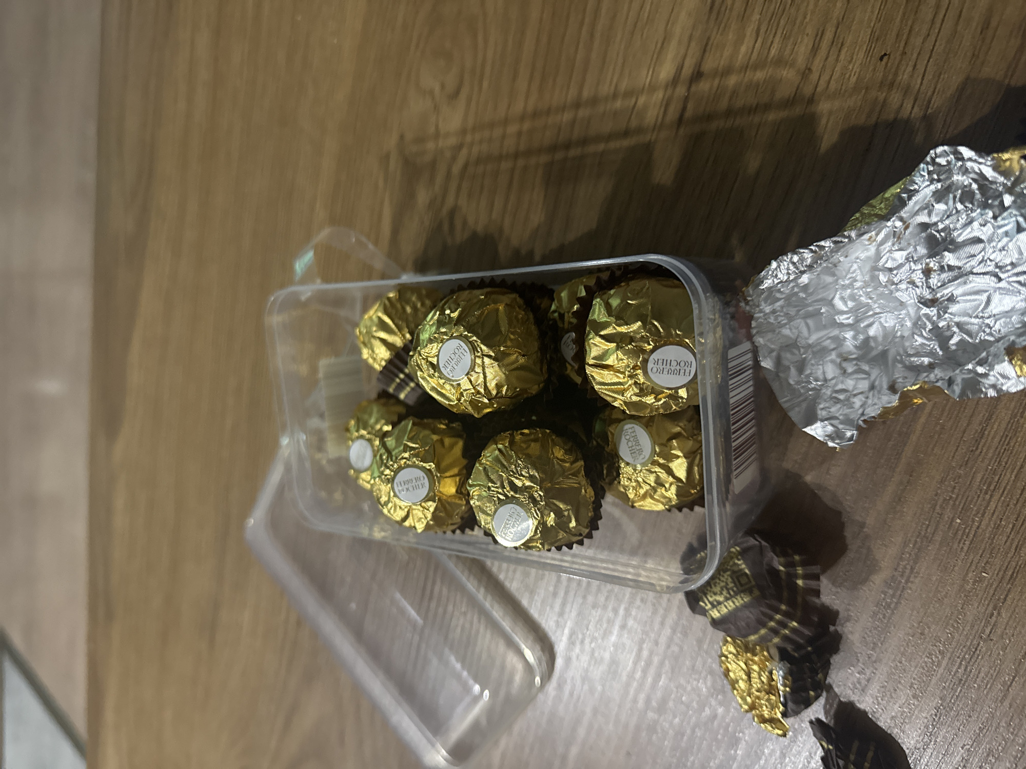 A clear plastic container holds seven individually wrapped Ferrero Rocher chocolates. The chocolates are spherical and wrapped in gold foil with a white circular sticker displaying the brand name. One chocolate at the bottom left of the container is partially unwrapped, revealing its brown wrapper and a portion of the gold foil. The container is positioned on a wooden surface, likely a table. To the left of the container, a crumpled brown and gold wrapper lies on the wooden surface. To the right of the container, a crumpled silver wrapper is also visible on the table. The background consists of a wooden surface with visible grain. The image does not contain any information that indicates a specific location in Tbilisi, Georgia.