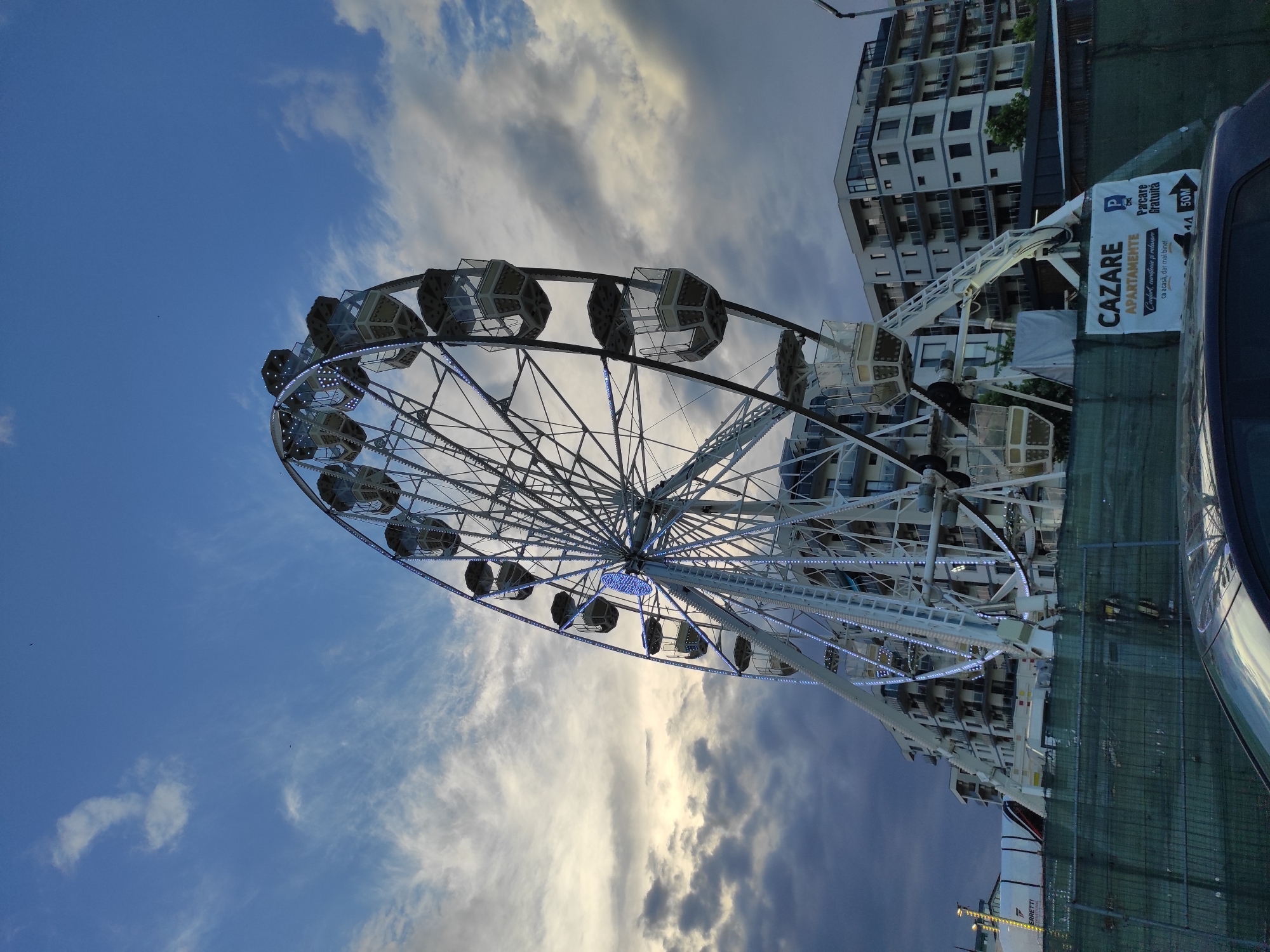 A low-angle, full shot captures a Ferris wheel situated in Năvodari, Romania, under a dynamic sky featuring blue patches and dramatic clouds. The Ferris wheel, illuminated by blue lights along its structure, dominates the center of the frame, with its passenger cabins visible. To the right of the Ferris wheel, a modern apartment building with multiple floors and balconies is depicted. In the foreground, a green construction fence is visible, with a car partially obstructing the bottom of the image. A sign attached to the fence reads "CAZARE APARTAMENTE" and "Parcare Gratuită" with an arrow indicating a distance of "50M". The overall impression is that of a seaside resort or developing area.