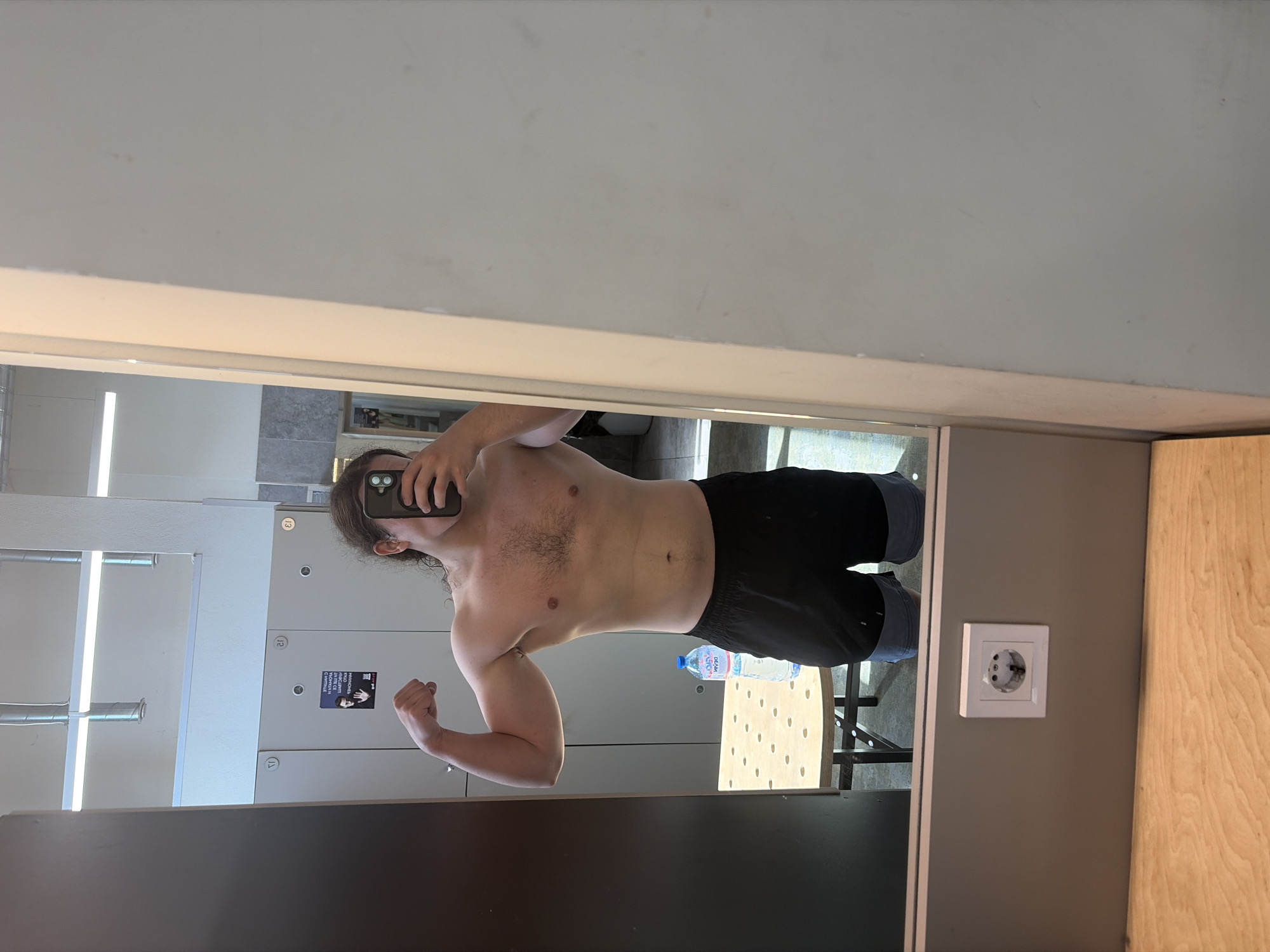 A shirtless man with long hair is posing in a mirror, flexing his arm in a gym-like locker room. The man is holding a phone in one hand and is taking a photo of himself. The background includes lockers, a water bottle, and a chair. The wall has an electrical outlet. The location is most likely in Plovdiv, Bulgaria, given the prompt.