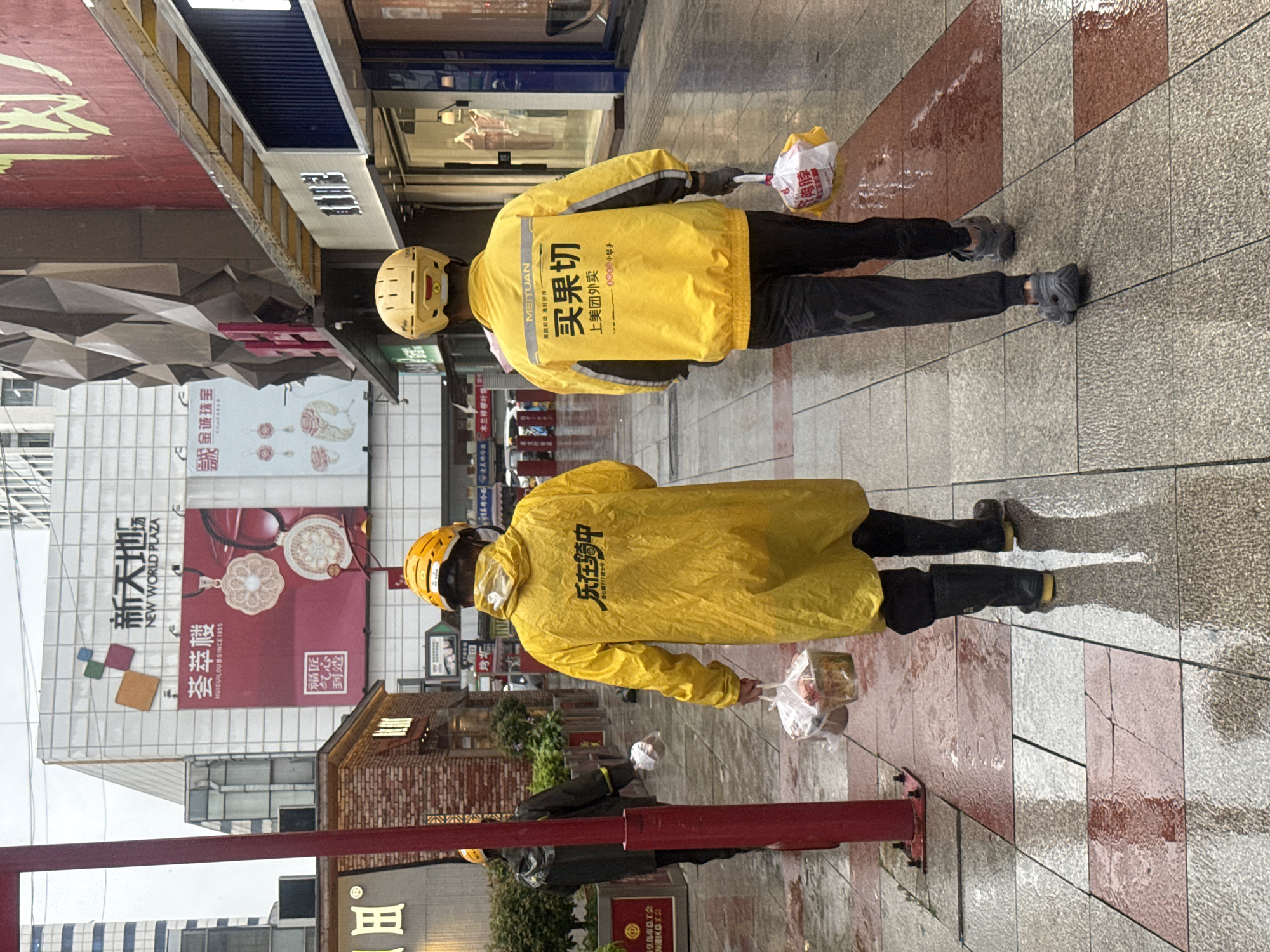 Two individuals, likely delivery workers, are walking away from the camera on a wet, tiled sidewalk in an urban environment. The person in the foreground wears a bright yellow raincoat with a white helmet, black pants, and black boots. They are carrying a clear plastic bag containing food items. The person behind them is also wearing a yellow raincoat and helmet, black pants, and grey athletic shoes, carrying a similar plastic bag. The background features a large building with signage that includes "NEW WORLD PLAZA" in English and Chinese. Storefronts with large windows are visible along the street. The ground is wet, indicating recent rain, with reflections of the surroundings visible on the surface. The overall scene suggests a busy commercial street in Qinhuangdao, China, during or after rainfall, with delivery personnel on duty.